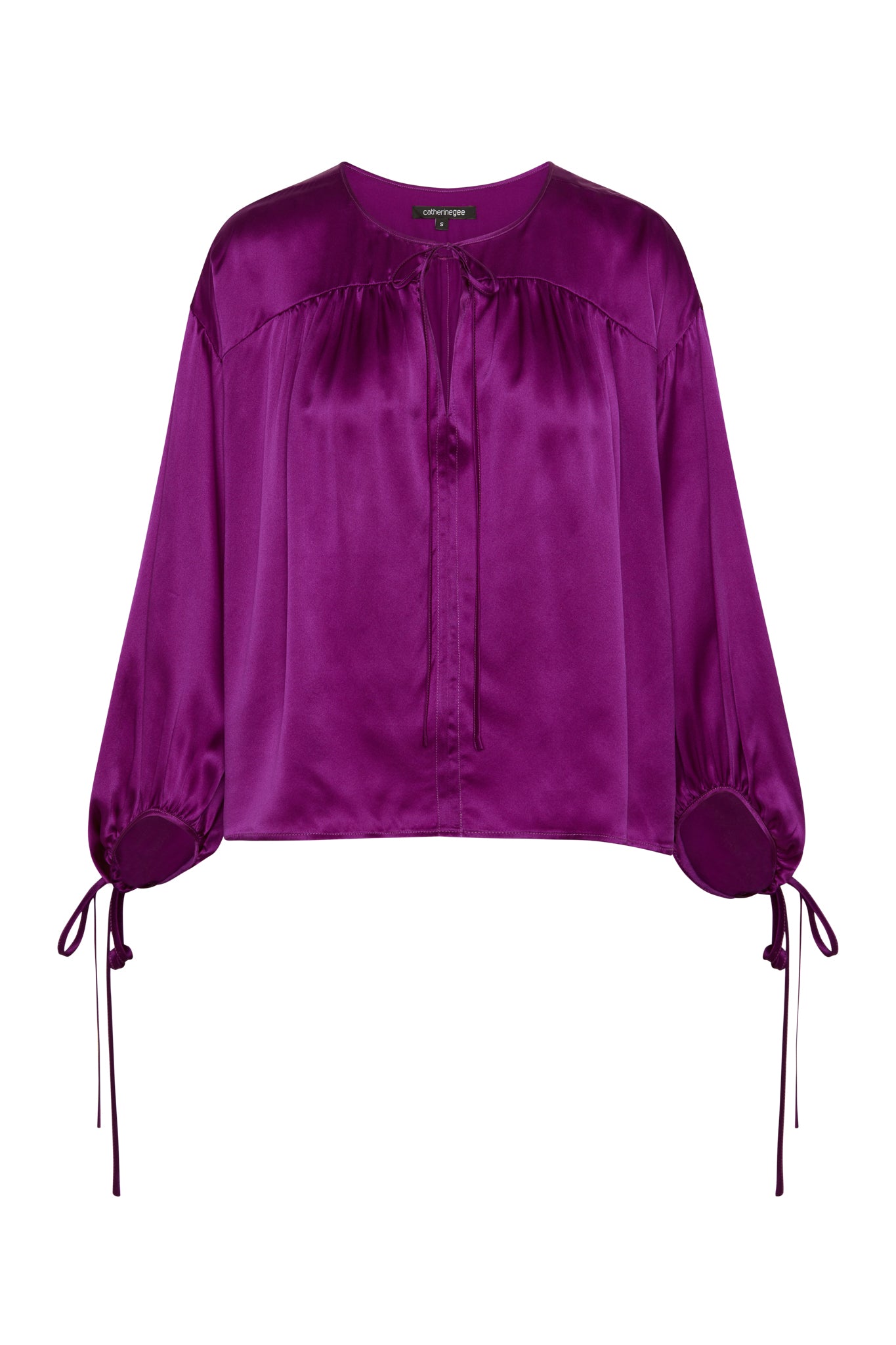 Catherine Gee Frances Electric Orchid Blouse is made of a luxurious, shiny fabric in a vibrant electric orchid color. It features a loose, flowing fit with long sleeves that gather at the wrists and are tied with delicate strings. The blouse has a round neckline with a subtle tie detail at the front, adding a touch of elegance to its overall design.