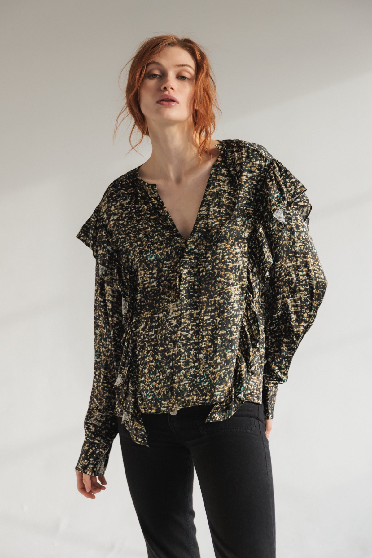 A woman wearing a Catherine Gee Bella - Gold Sequin blouse features a loose fit with a deep V-neckline and long sleeves paired with black pants. The fabric has a speckled gold sequin pattern, creating a shimmering effect.
