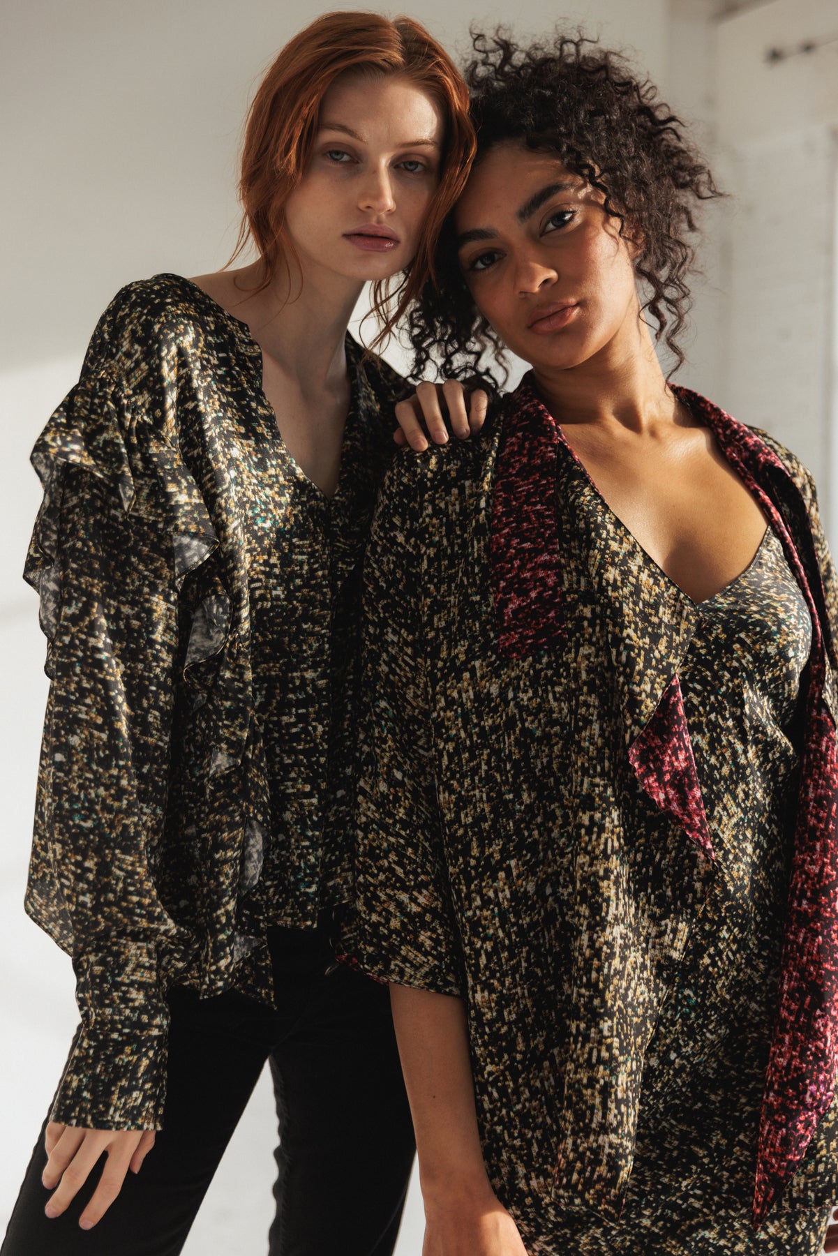 Two models are wearing Catherine Gee Bella Blouse - Gold Sequin. The blouse on the left features long sleeves with ruffle details and a shimmering gold sequin pattern. The model on the right pairs the blouse with a matching sequin top underneath and a contrasting red scarf. The image showcases the elegant and stylish design of the blouses, emphasizing their textured, shiny fabric.