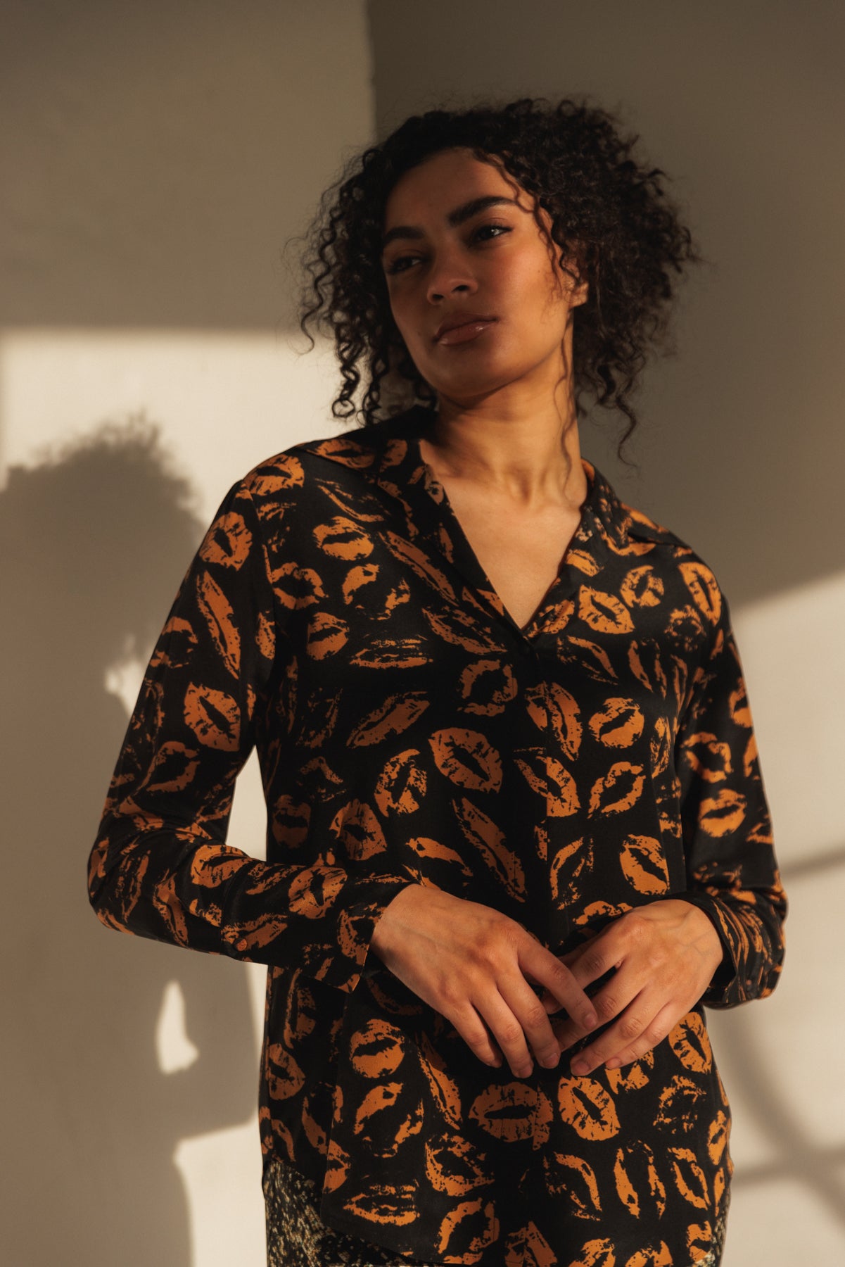 A woman with her hands positioned in front of her stomach, with fingers interlaced standing against a wall with a shadow cast behind her wears a Catherine Gee Daria French Cuff Silk-Mocha Lips blouse that features a black background and a mocha-colored lip print pattern long-sleeved. The lighting in the image creates a warm and soft atmosphere, highlighting the texture and pattern of the blouse.