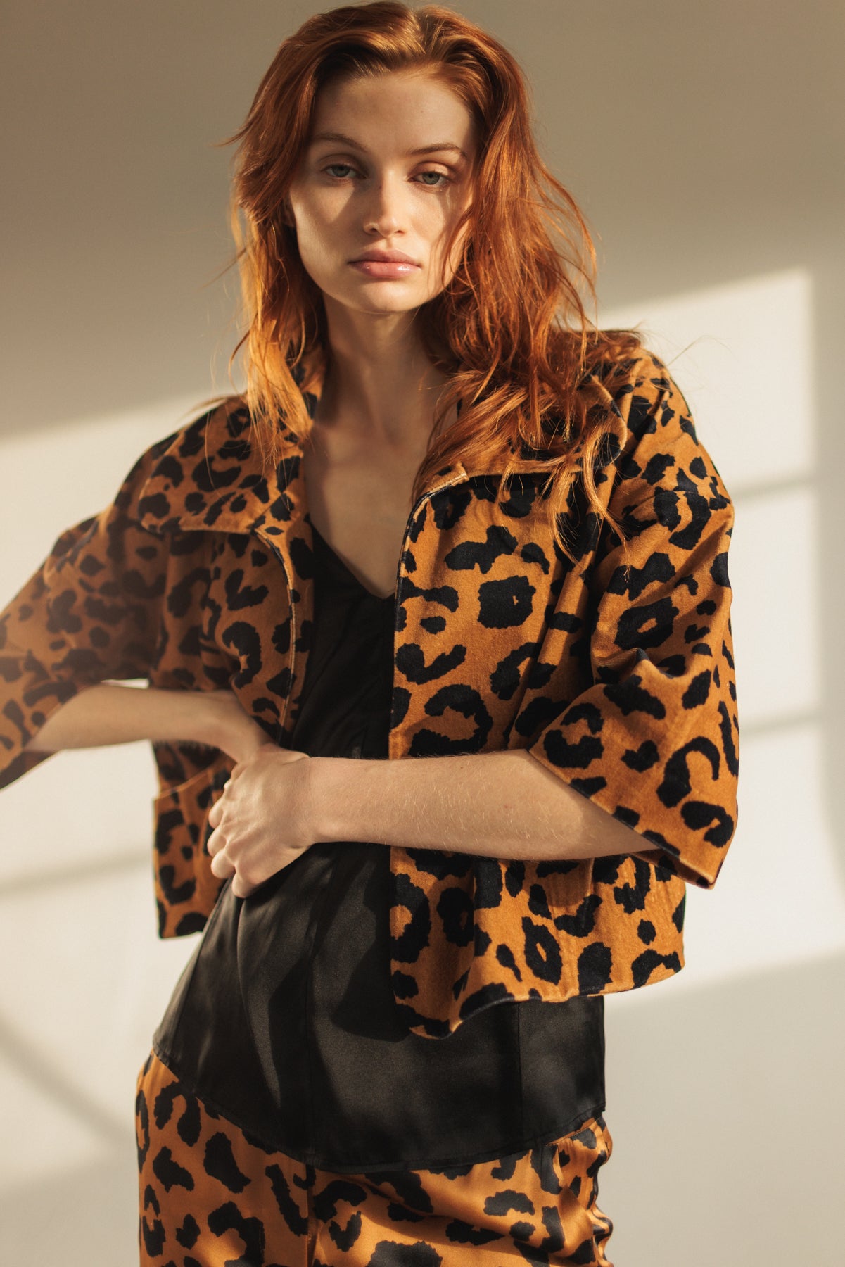 A woman is wearing a Catherine Gee Alexa Cropped Jacket in a cheetah print that features a bold cheetah pattern in shades of brown and black paired with cheetah print pants and a black top underneath the jacket. The image is well-lit with natural light, highlighting the texture and pattern of the clothing.
