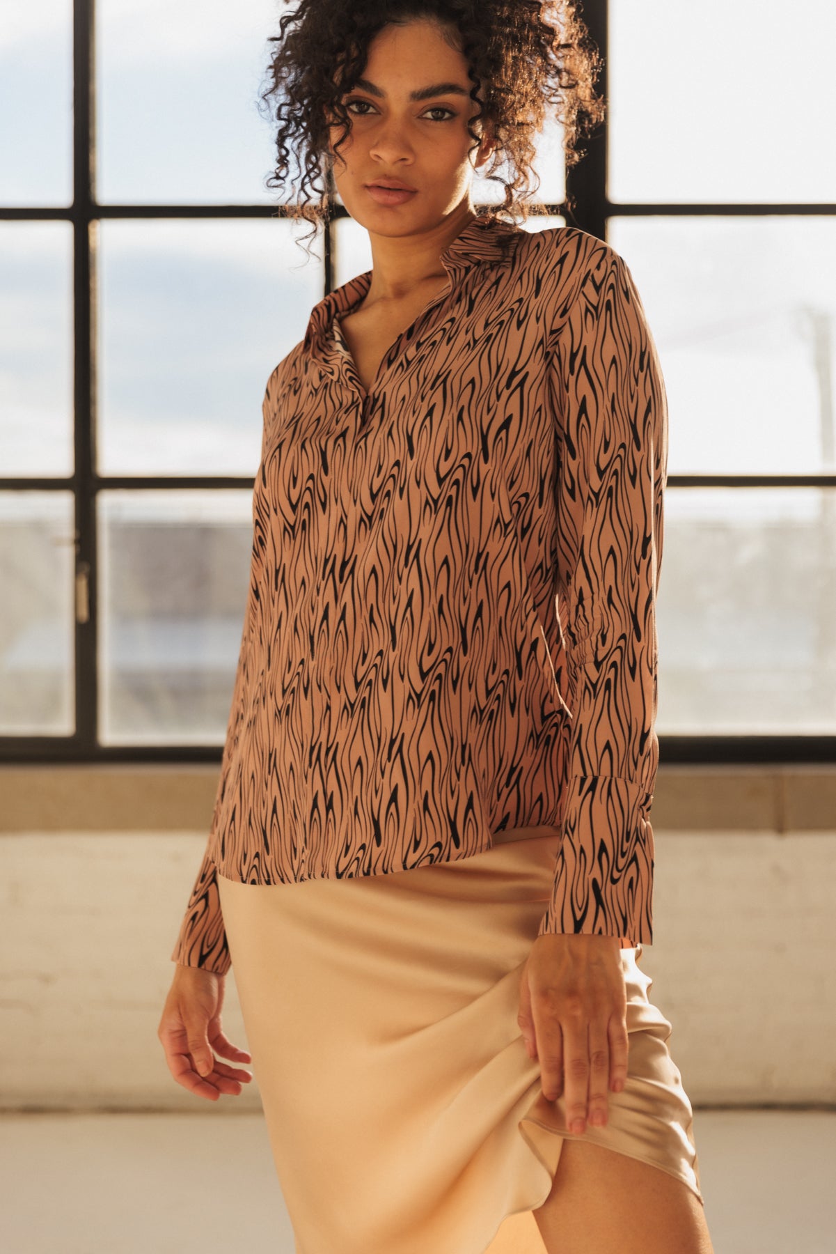 A woman wearing the Catherine Gee Daria French Cuff Silk Wishbone Blouse. This elegant blouse features a neutral wishbone pattern, with long sleeves and a relaxed fit paired with a light-colored skirt, creating a sophisticated look. The background of large windows letting in natural light adds a soft and elegant ambiance, highlighting the blouse's stylish design.