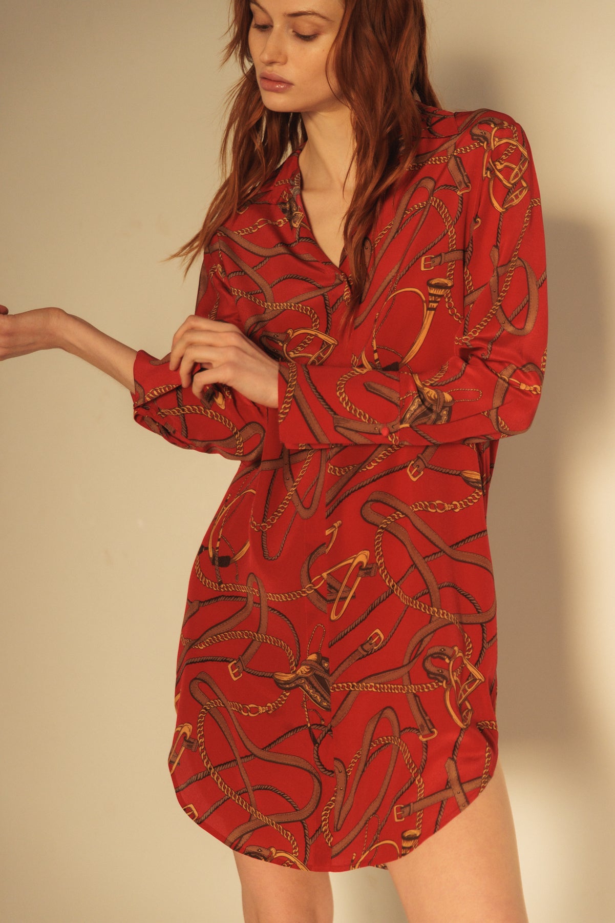 A woman wearing a Catherine Gee Daria Silk Shirt Palermo dress is primarily red in color with a pattern of intertwined ropes and chains in shades of gold and brown. It has long sleeves and a V-neckline, giving it a smooth and elegant appearance. The fabric looks silky and luxurious, making this stylish and eye-catching dress perfect for fashion enthusiasts.