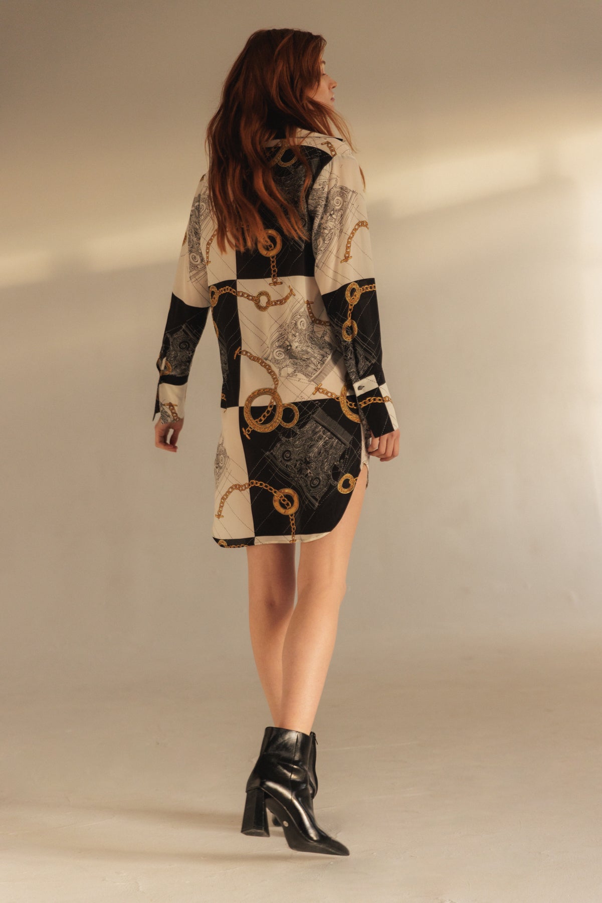 A woman wearing a Catherine Gee Daria Silk Shirt Athens dress features a black and white pattern with gold chain designs and complex motifs. She is standing with her back to the camera, showcasing the dress's long sleeves and above-the-knee length paired with black heeled ankle boots, adding a stylish touch to the overall look in a neutral background, highlighting the dress's design.