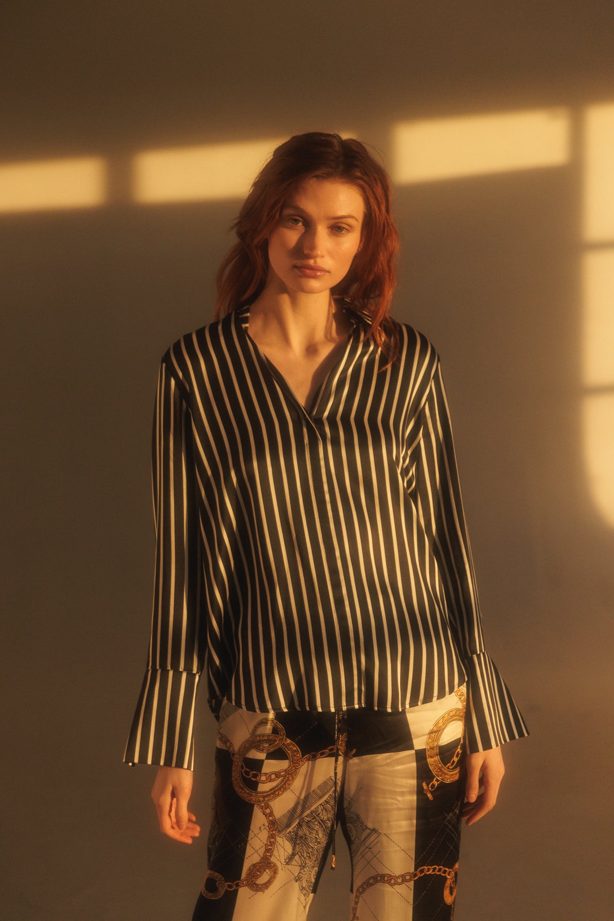 A woman wearing a Catherine Gee Daria French Cuff Silk black and white stripes blouse features long sleeves with French cuffs and a relaxed fit. She is also wearing patterned pants with chain and rope designs. The image is taken in warm lighting, casting soft shadows on the wall behind, highlighting the stylish and sophisticated design of the blouse.