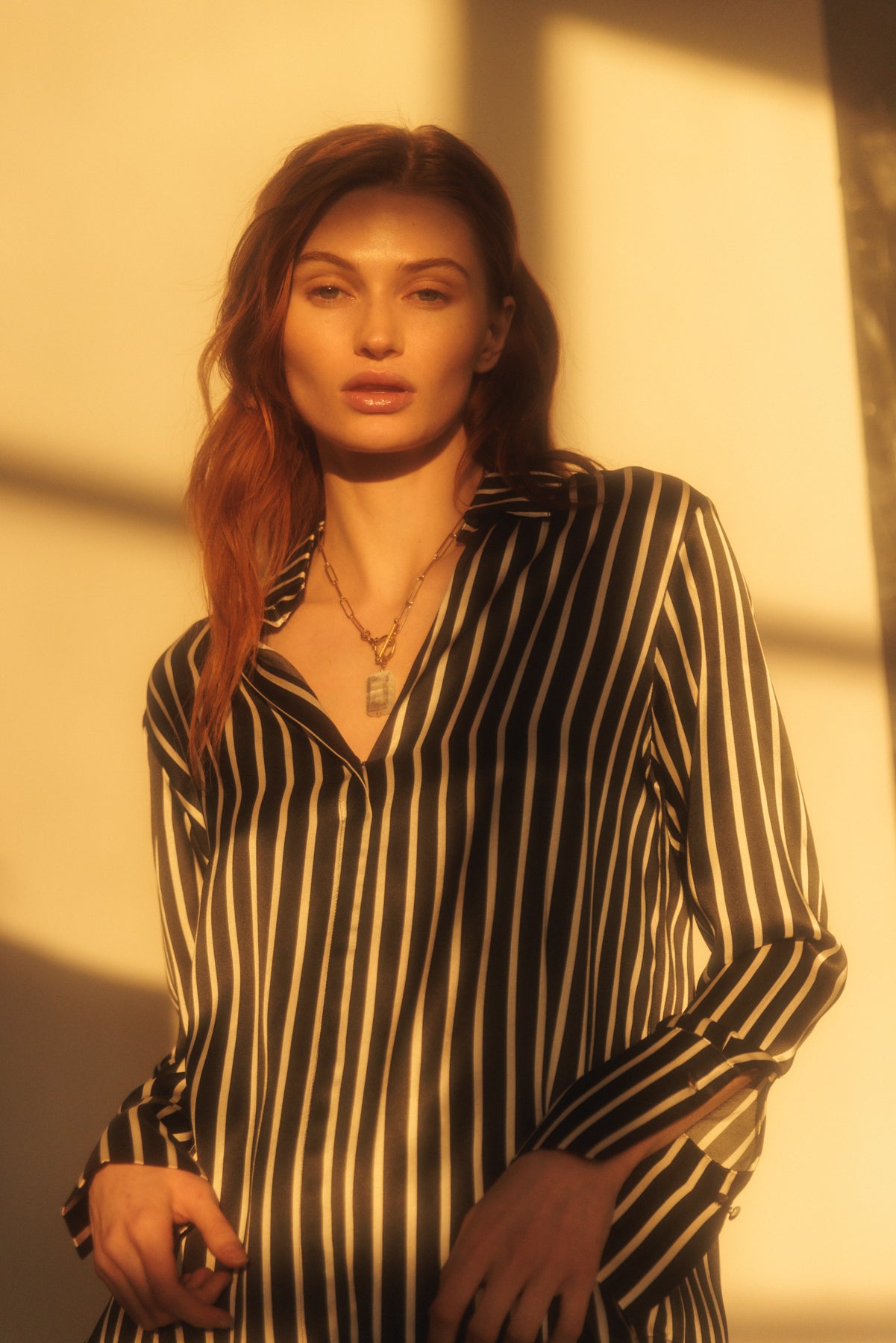 A woman wearing a Catherine Gee Daria French Cuff Silk black and white stripes blouse features a deep V-neckline and long sleeves with French cuffs. She is also wearing a necklace with a rectangular pendant. The lighting in the image creates a warm, golden hue, adding a soft and elegant ambiance to the scene.