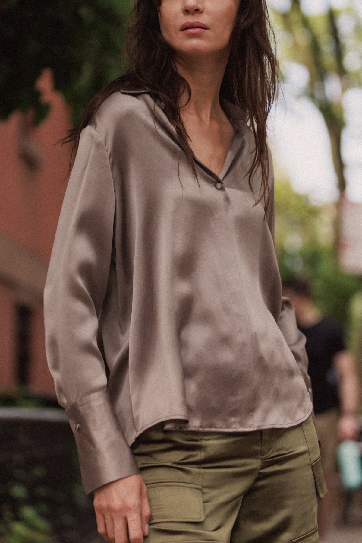 Woman wearing a Catherine Gee Daria French Cuff Silk-Grey blouse made of smooth, shiny silk fabric and features a relaxed fit with a V-neckline and French cuffs paired with olive green cargo pants. The background is an outdoor setting with blurred greenery and buildings.
