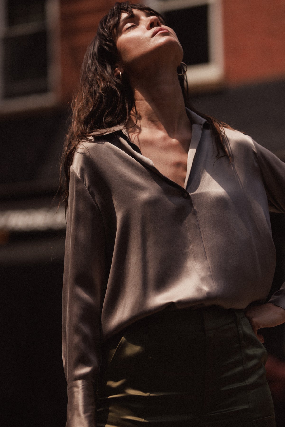 A woman wearing a Catherine Gee Daria French Cuff Silk-Grey blouse made of smooth, shiny silk fabric and features a deep V-neckline with a relaxed fit paired with high-waisted pants in a dark color. The background shows an urban setting with blurred buildings highlighting the elegant and luxurious texture of the silk blouse.
