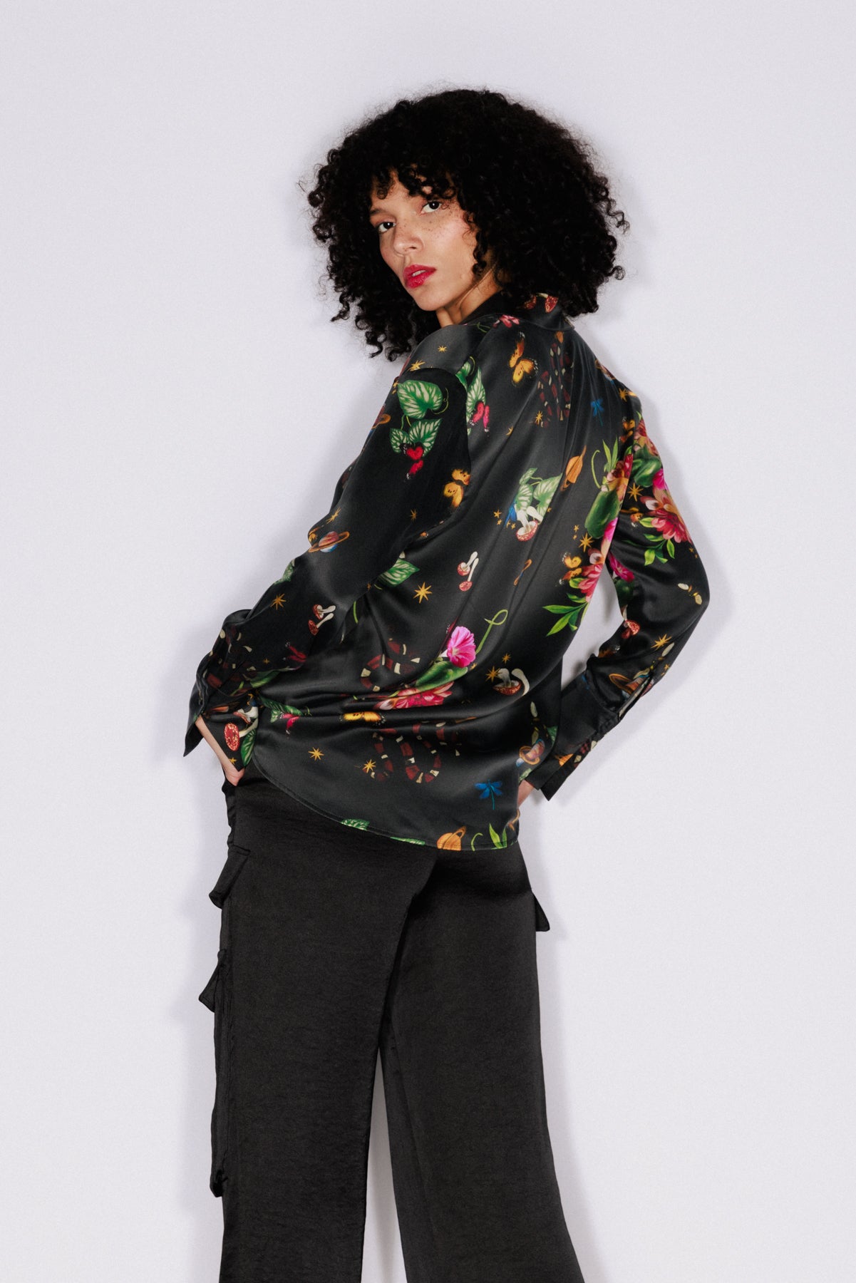 A woman wearing a Catherine Gee Daria French Cuff Silk Serpentine blouse features a black base color with a vibrant, eclectic print featuring various elements such as flowers, mushrooms, snakes, butterflies, and planets paired with dark-colored pants. The fabric appears to be silk, giving it a smooth and shiny texture, and has long sleeves with French cuffs, adding a sophisticated touch to the overall design.