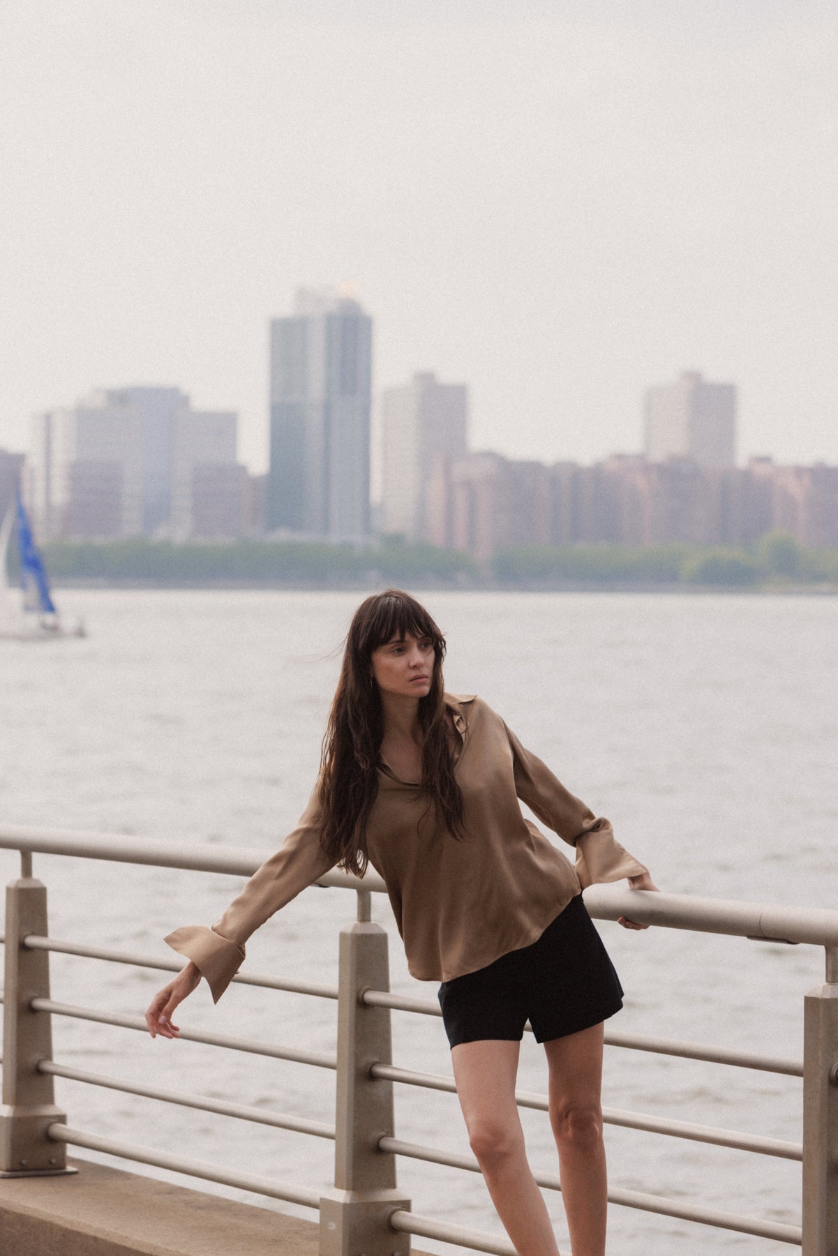 A woman with long dark hair standing by a railing near a waterfront in the background of a city skyline with high-rise buildings in a calm atmosphere in a late afternoon scene. She wears a Catherine Gee Daria French Cuff Silk-Mocha blouse featuring a loose brown top and black shorts, leaning slightly to one side, with one hand resting on the railing. A sailboat can be seen in the distance on the water giving a serene and relaxed vibe.