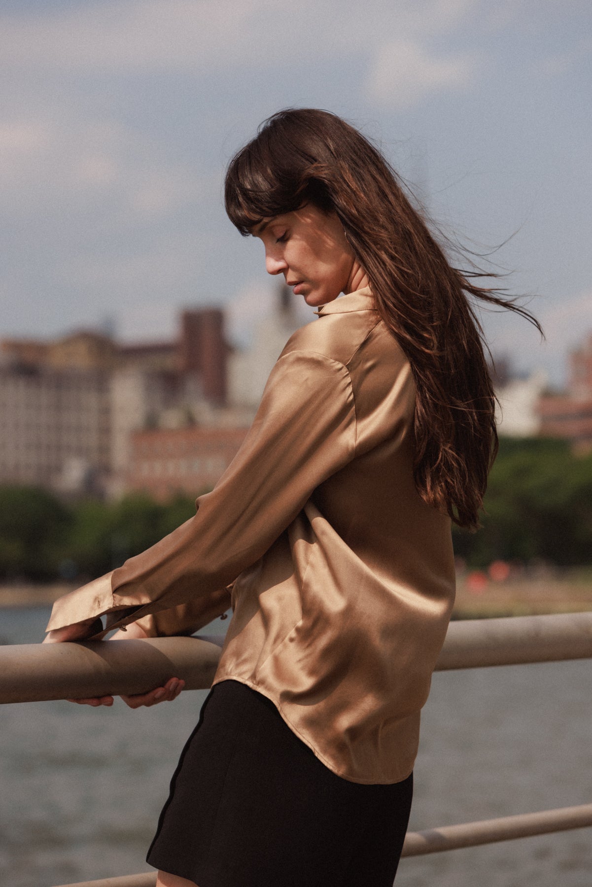 A woman standing outdoors near a railing, with her head slightly tilted down and to the side, giving the impression of reflection. She is wearing a Catherine Gee Daria French Cuff Silk-Mocha blouse featuring a satin-like mocha shirt, and the wind seems to be gently blowing her long dark hair. The background features a cityscape with buildings and trees, creating a warm, calm, and reflective atmosphere, adding a serene and peaceful vibe.