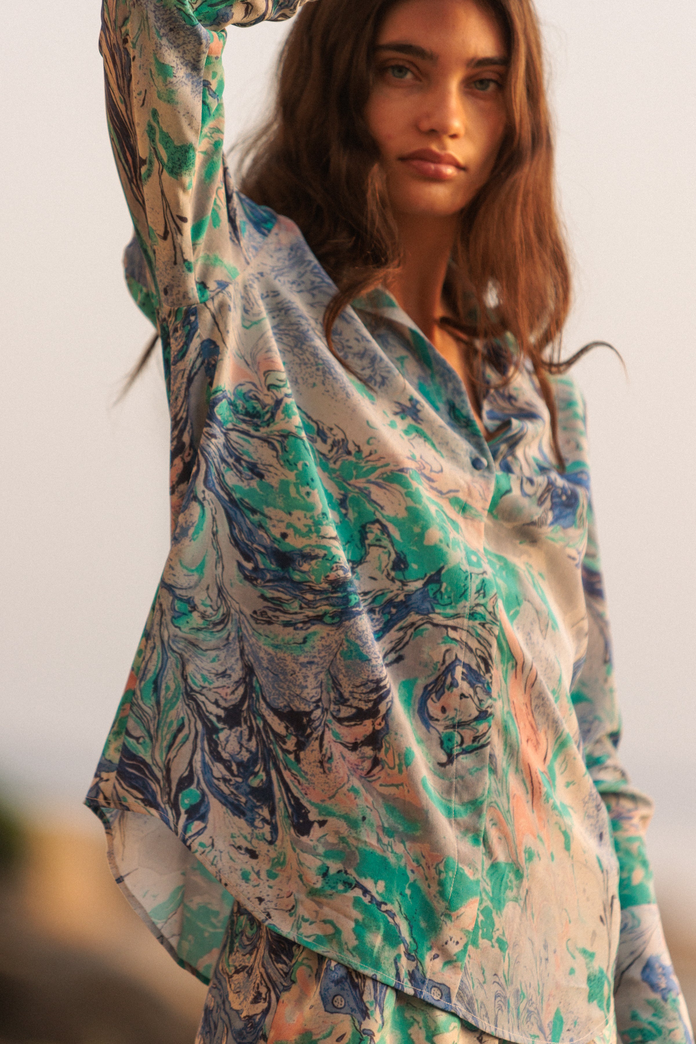 A woman wearing a Catherine Gee Daria French Cuff Silk Cool Swirl blouse features a vibrant, abstract design with shades of blue, teal, and light peach, resembling a marbled effect posing with one arm raised highlighting the blouse's loose fit and elegant drape. The fabric appears lightweight and flowy.