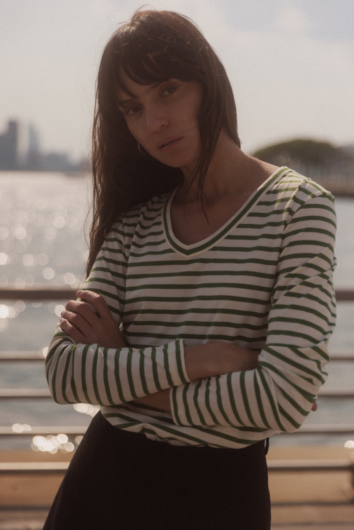 A woman is standing outdoors near a waterfront, with her arms crossed and leaning against a railing. The background features a blurred view of the sea and a distant cityscape. She is wearing a Catherine Gee Jen Relaxed Longsleeve in a green stripe pattern. The tee has green and white horizontal stripes, a V-neckline, and long sleeves. The lighting indicates a sunny day, with reflections on the water creating a serene ambiance for the scene.