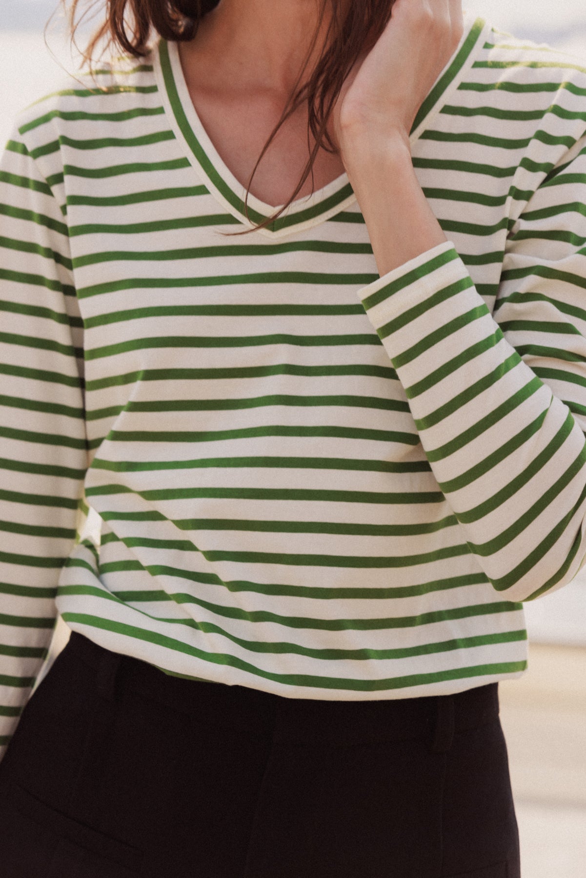 A woman wearing a Catherine Gee Jen Relaxed Longsleeve - Green Stripe Tee. The tee features horizontal green stripes on a white background, with a V-neckline and long sleeves paired with black pants. The relaxed fit and classic stripe pattern make this tee a versatile and stylish choice for casual wear.