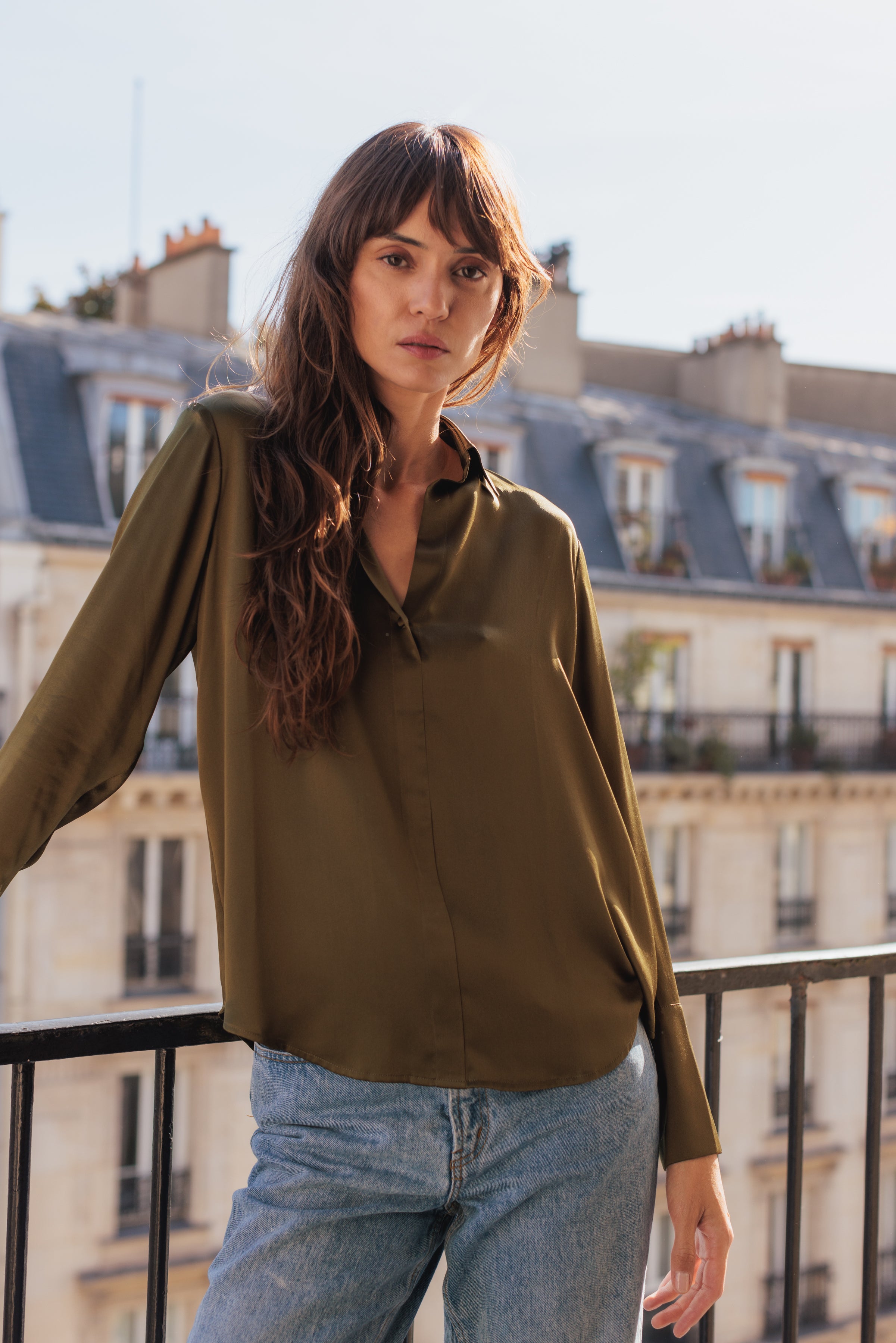 A woman standing on a balcony wearing a Catherine Gee Daria French Cuff Silk Blouse Army Green, paired with light blue jeans. The backdrop features classic Parisian-style buildings, highlighting a casual yet stylish urban look.