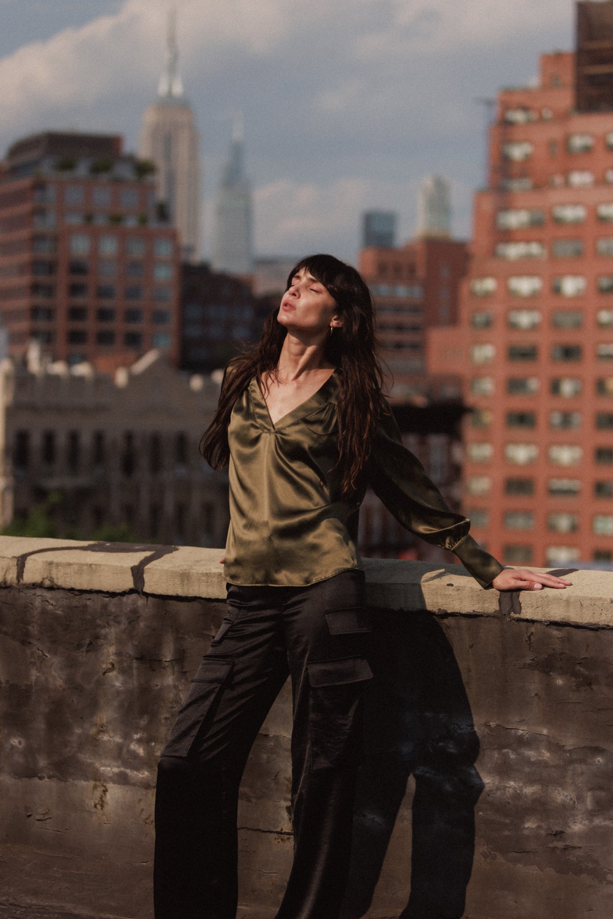 A woman is standing on a rooftop wearing Catherine Gee's black cargo pants paired with a silky olive green blouse. The background shows a cityscape with various buildings, adding an urban feel.