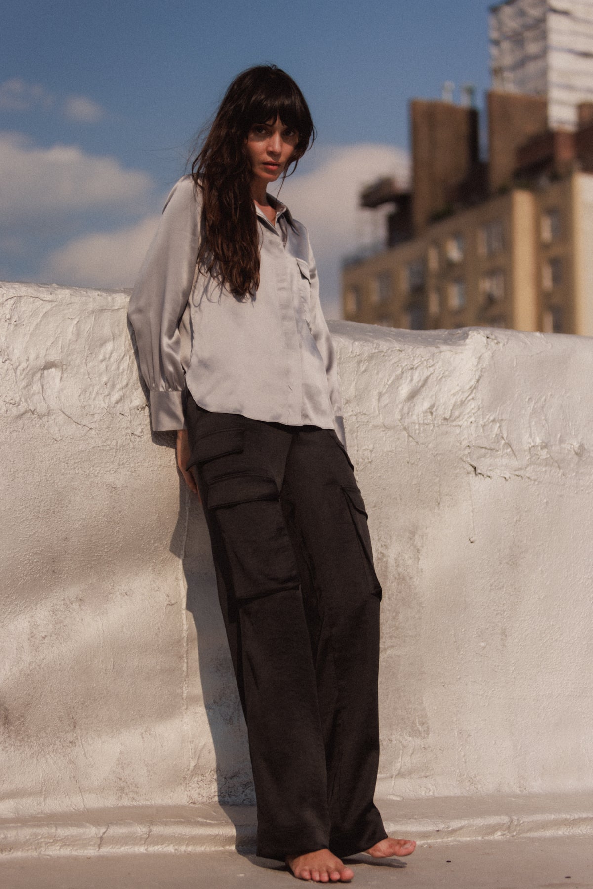 A woman is standing on a rooftop against a white wall, wearing a light-colored long-sleeve shirt and Catherine Gee's black cargo pants. The background shows a clear sky with some clouds and buildings in the distance highlighting the black cargo pants, which have multiple pockets and a loose fit.