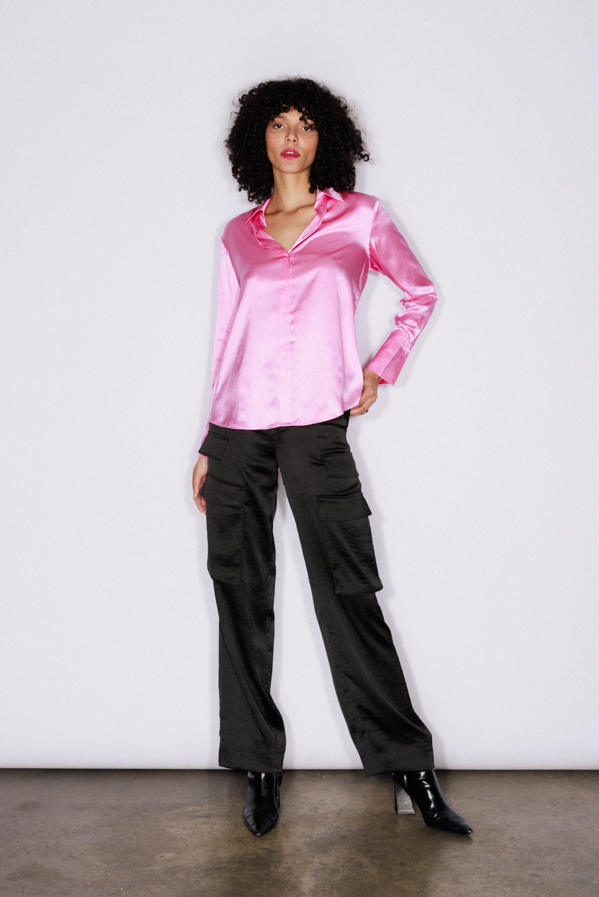 A woman wearing a Catherine Gee Daria French Cuff Silk Doll Pink blouse features a shiny, smooth texture with a classic collar and long sleeves ending in French cuffs paired with black cargo pants with multiple pockets and black pointed-toe ankle boots. The background is plain white, and she is standing on a concrete floor.