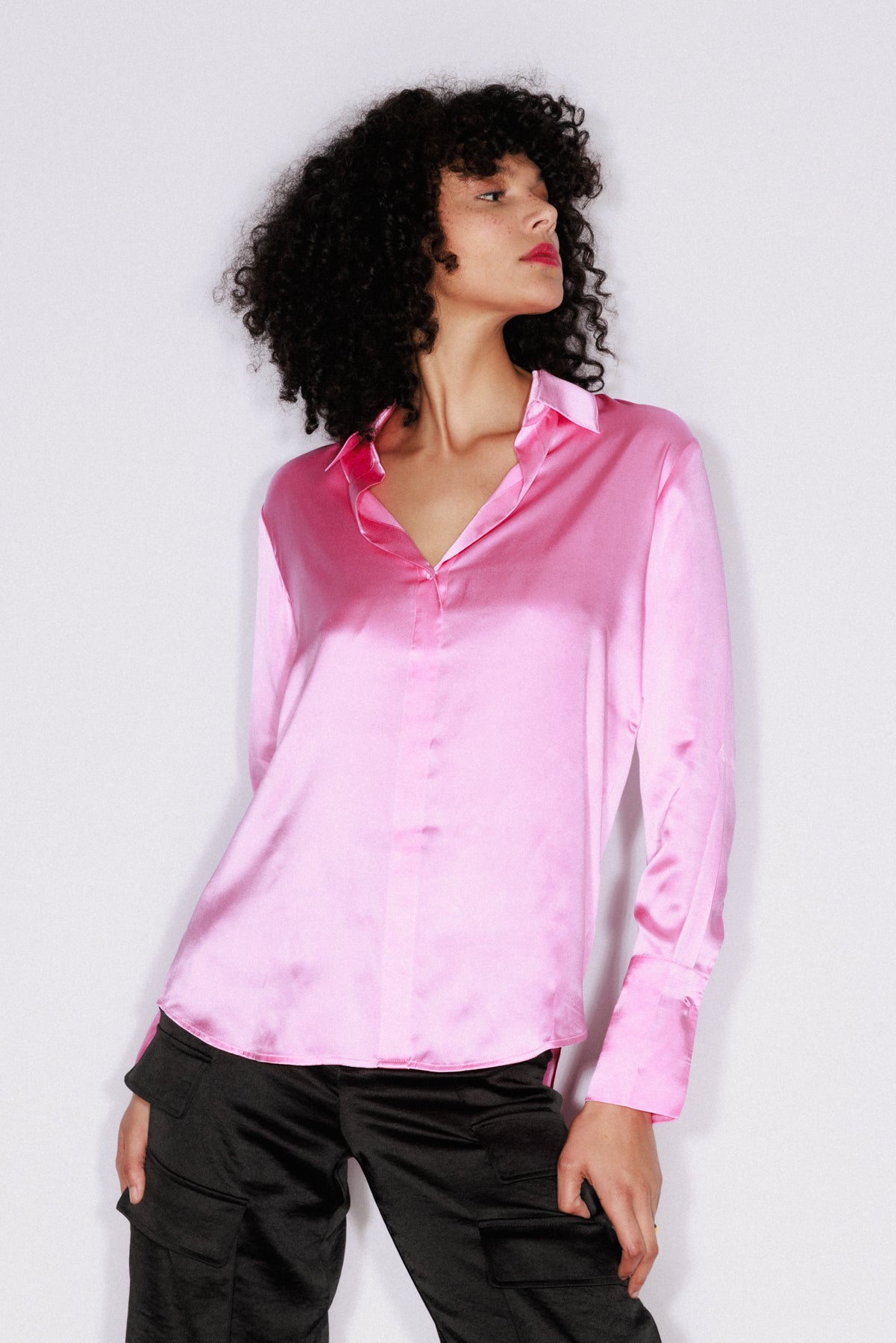 Woman wearing a Catherine Gee Daria French Cuff Silk Blouse in Doll Pink. The blouse is made of shiny silk fabric, has a classic collar, and long sleeves with French cuffs. She is also wearing black pants with pockets on a plain white background.