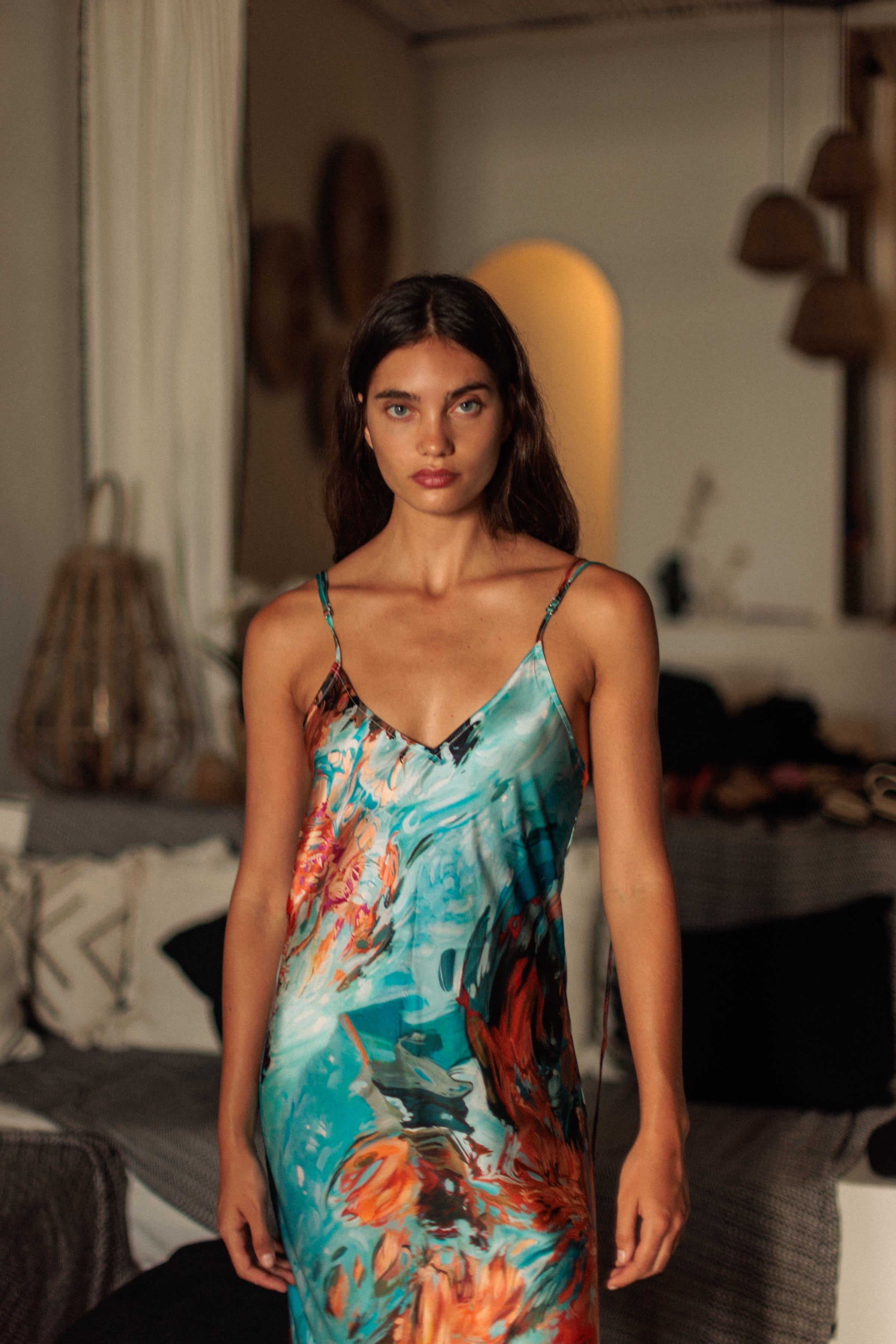 A woman wearing the Catherine Gee Emma 7 Girls Slip dress showcases a vibrant and artistic print featuring a blend of turquoise, orange, and black, creating a visually striking pattern. The dress is designed with thin spaghetti straps and a V-neckline, giving it a delicate and elegant look. The background includes soft lighting and home decor elements, adding a cozy and intimate atmosphere that highlights the beauty of the dress.