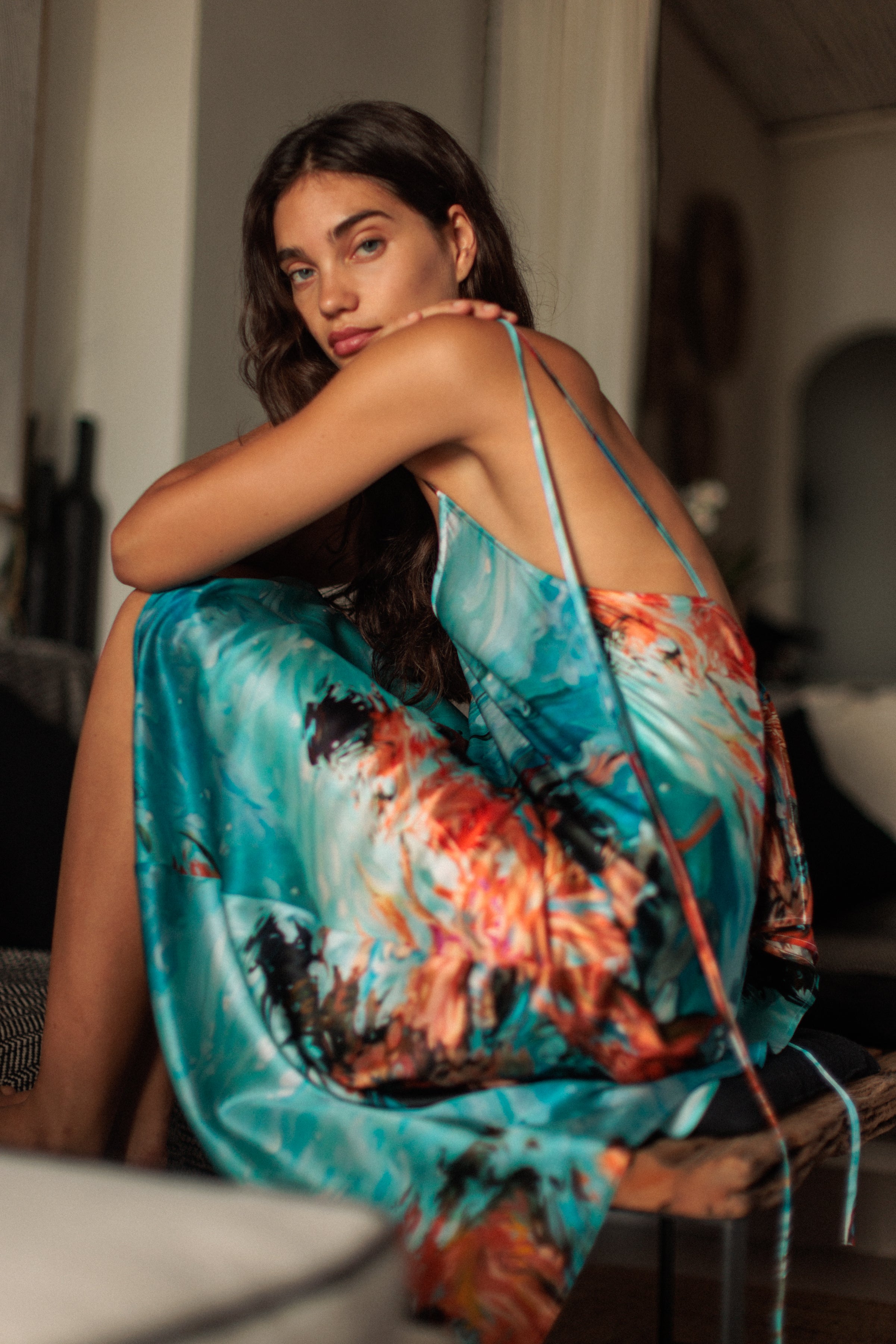 A woman sitting with knees drawn up, wearing a Catherine Gee Emma 7 Girls Slip dress features a vibrant, abstract pattern with a mix of turquoise, orange, and black colors. The dress has thin straps and an open back, giving it a delicate and elegant appearance. The blurred background includes some furniture and decor elements, adding to the cozy and intimate atmosphere.