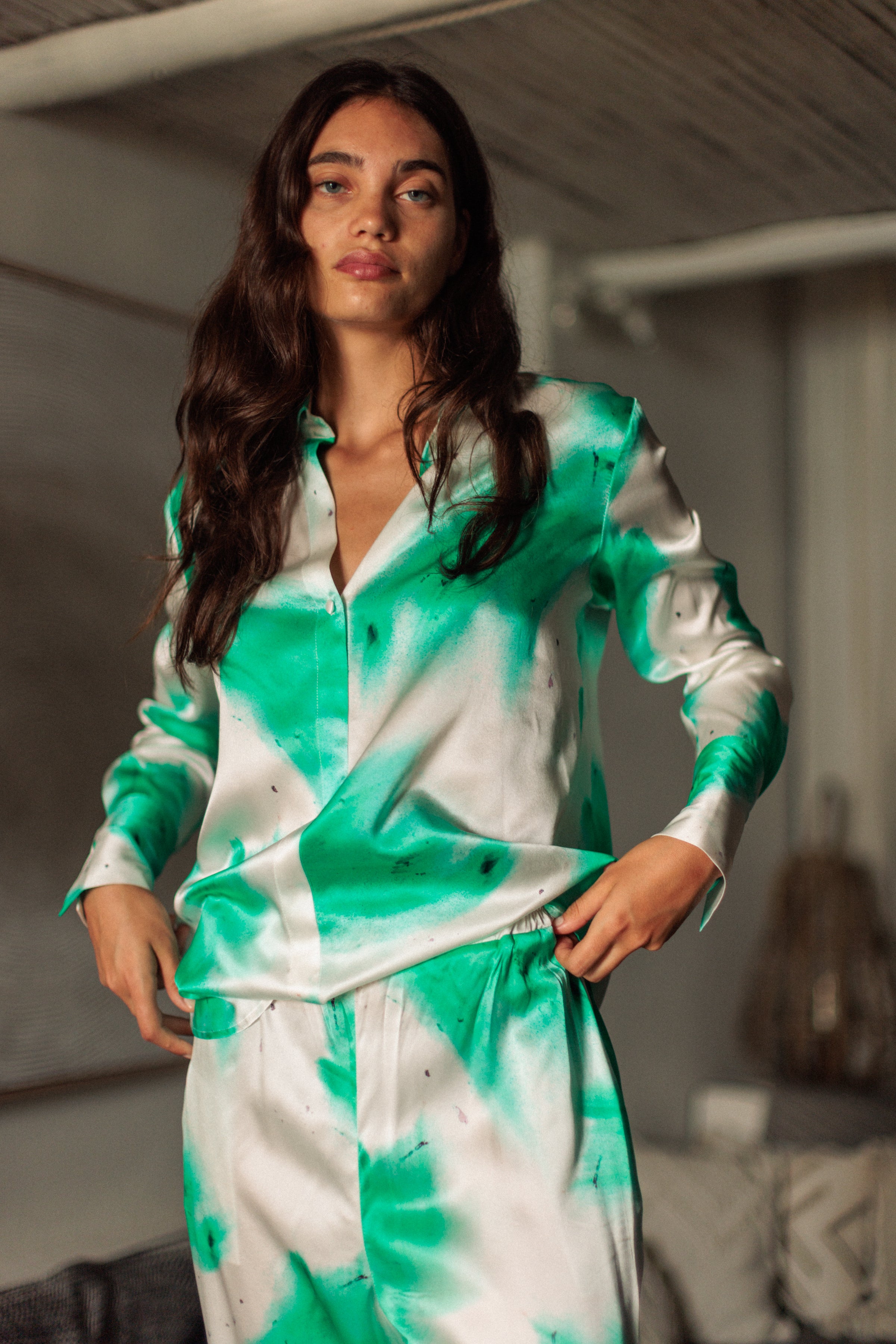 A woman wearing a Catherine Gee Daria French Cuff Silk Teal Art blouse features a vibrant teal and white abstract pattern, with a button-up front and French cuffs. The silk fabric gives it a luxurious and smooth appearance. She also wears matching pants with the same teal and white abstract design, creating a coordinated and stylish outfit in a cozy and modern background.