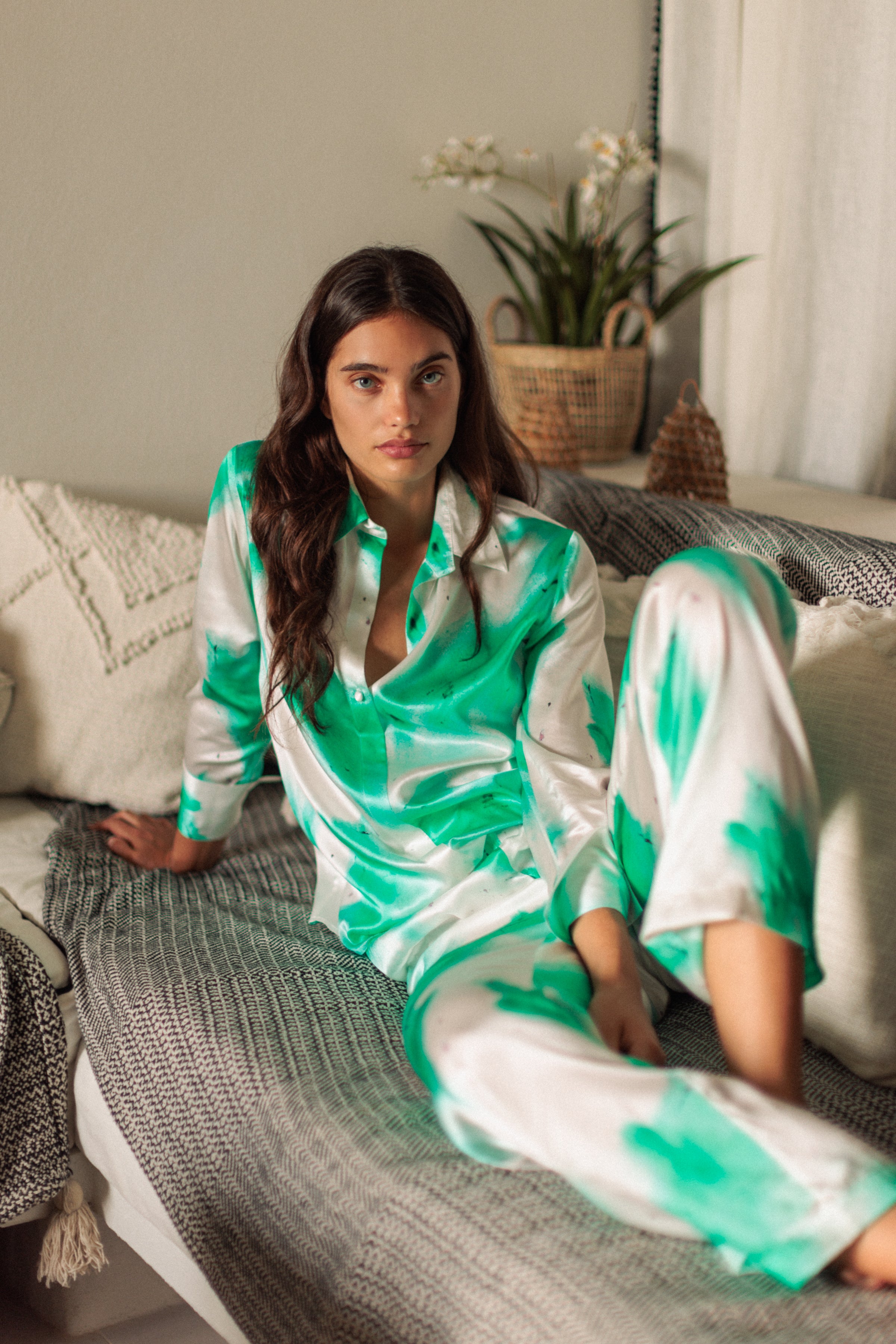 A woman sitting on a couch, wearing a Catherine Gee Daria French Cuff Silk Teal Art blouse and matching pants, features a vibrant teal and white abstract pattern, giving it a stylish and artistic look. The setting appears cozy, with a woven basket and plants in the background, adding to the relaxed atmosphere.