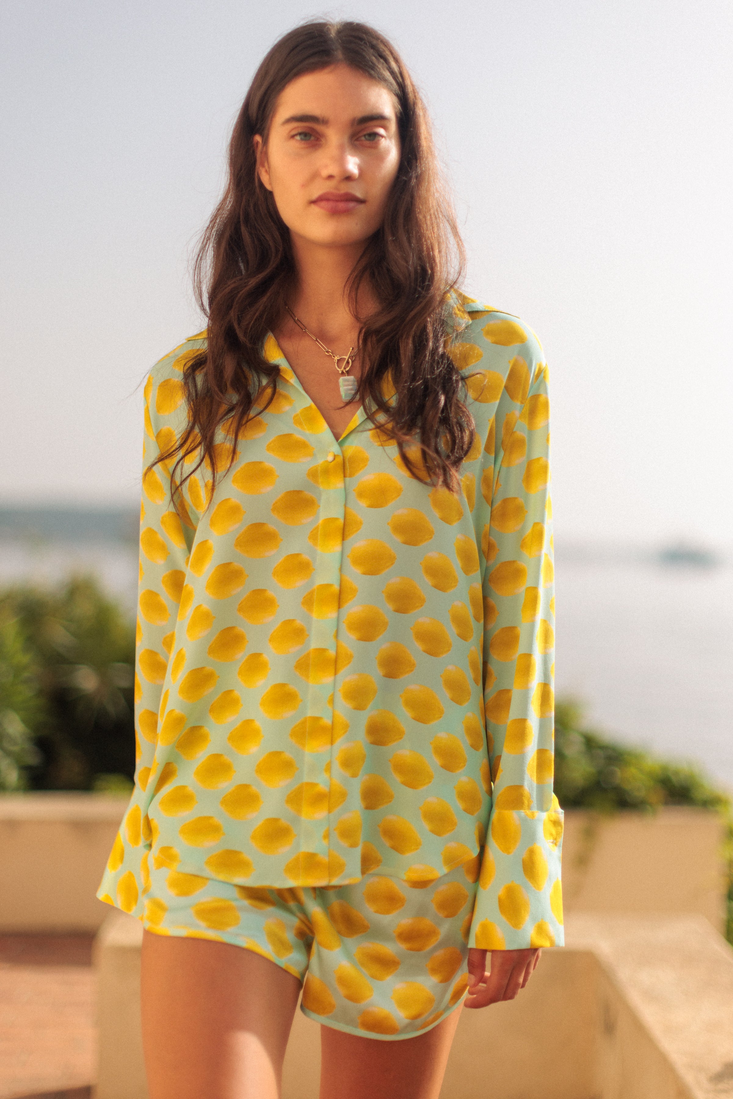 A woman wearing a Catherine Gee Daria French Cuff Silk-Limon Blue blouse features a light blue background with a playful pattern of yellow lemon-like spots. It has a classic collar and long sleeves with French cuffs. She is standing outdoors with greenery and a body of water in the background, suggesting a warm and sunny environment.
