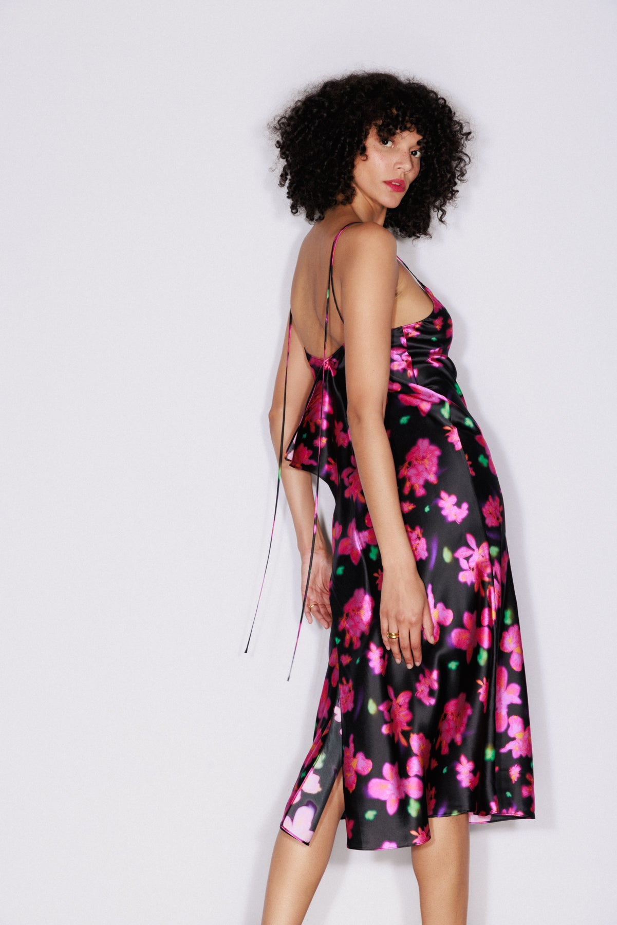 A woman standing sideways wearing the Catherine Gee Emma Neon Flower Slip Dress. This mid-length dress features a vibrant floral pattern with neon pink flowers on a black background, has thin spaghetti straps, and an open back with a tie detail. The fabric appears to be silky and smooth, giving it a luxurious and elegant look highlighting the dress's flowy and flattering fit.