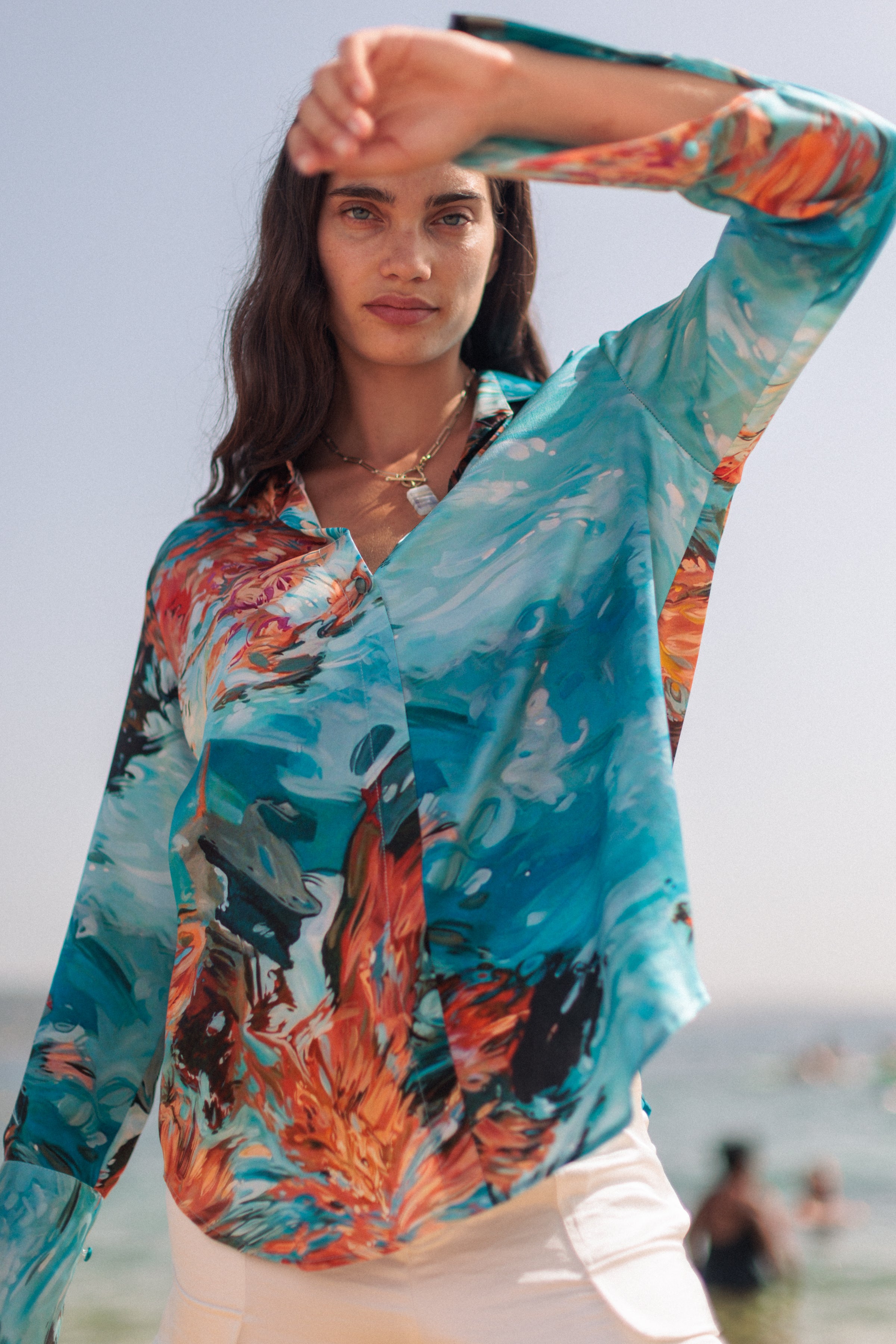 Woman wearing a Catherine Gee Daria French 7 Girls - Cuff Silk Blouse with vibrant, abstract-patterned silk with a mix of blue, brown, black, and orange colors. The blouse has a French cuff design and is paired with white pants. The background shows a beach scene with a clear sky and people in the distance.