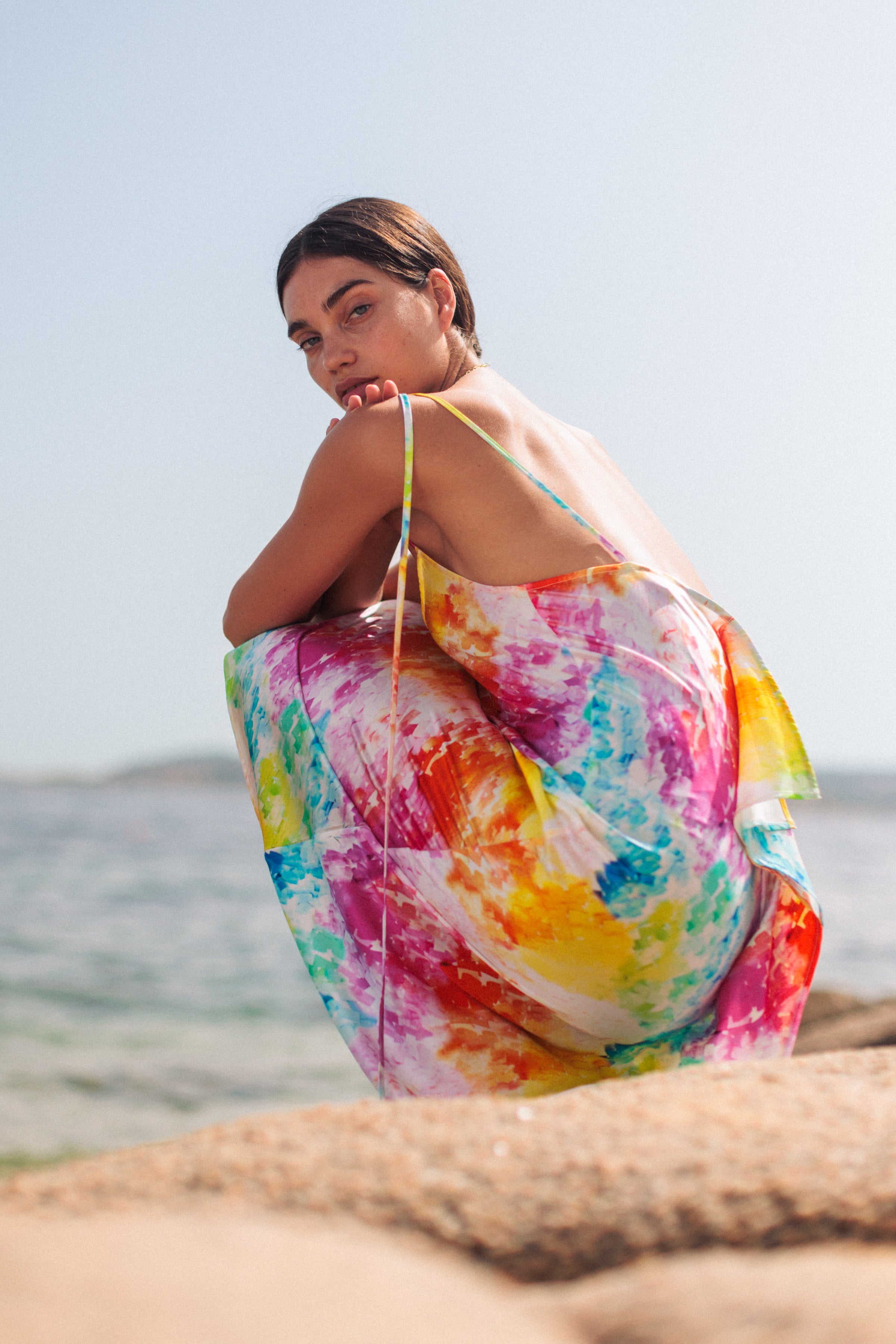 A woman wearing the Catherine Gee Emma Rainbow Moodboard Slip vibrant dress features a multicolored pattern with splashes of red, yellow, orange, pink, blue, and green. She is crouching on a sandy beach with the ocean in the background, creating a serene and picturesque scene. The dress's light and airy fabric, combined with the colorful design, makes it stand out against the natural backdrop.