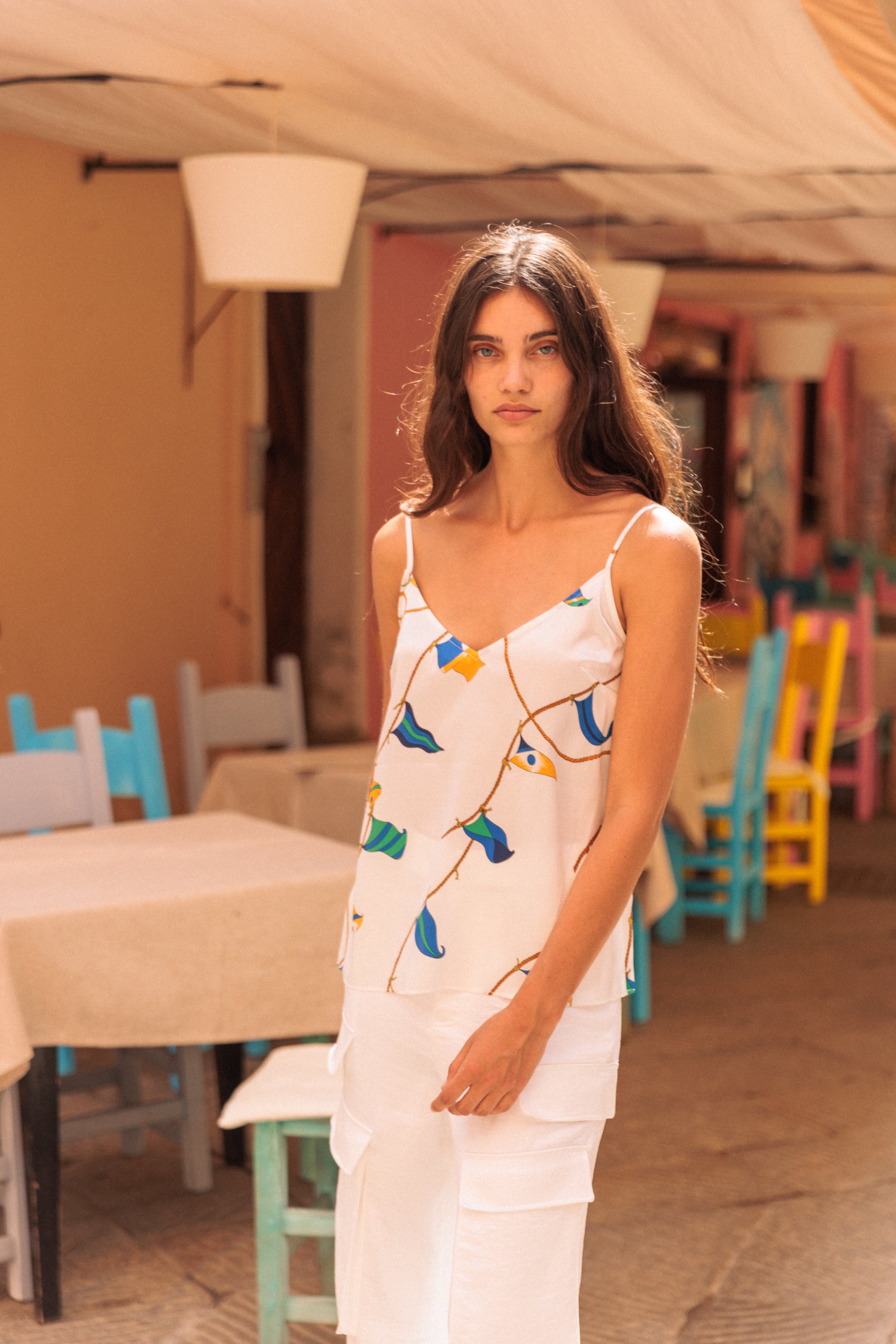 A woman wearing a Catherine Gee Franka Lisbon Sails cami features a white base with a colorful pattern of blue, green, and yellow sail-like shapes connected by thin, golden lines. She is standing in an outdoor setting with pastel-colored chairs and tables in the background, suggesting a casual, summery atmosphere. The cami's design is elegant and vibrant, making it a standout piece for warm weather.