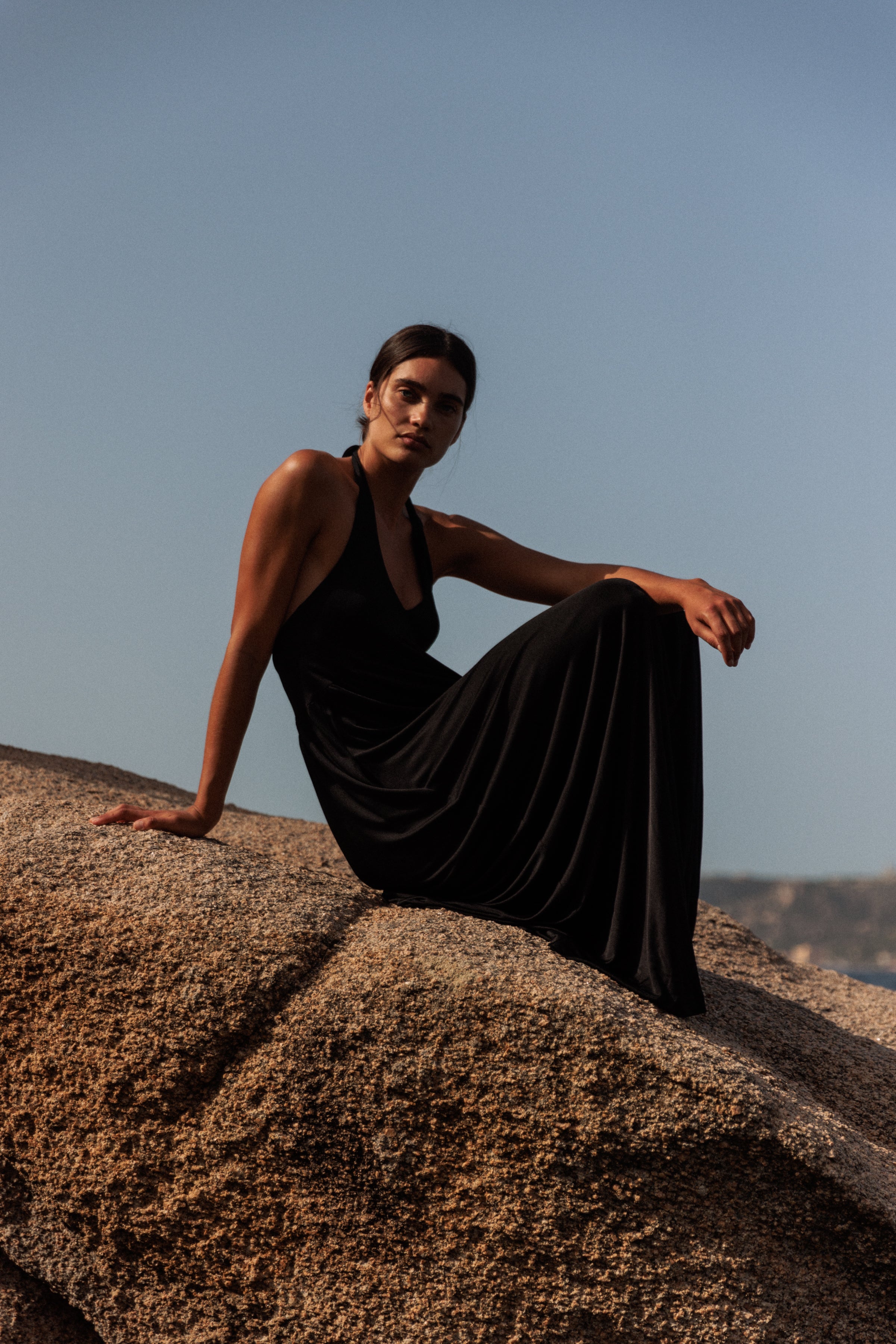 A woman sitting on a large rock outdoors, wearing a long, Anna Halter black dress by Catherine Gee. The background shows a clear sky and a distant landscape, suggesting a serene and natural setting, highlighting the elegance and flow of the dress, and making it relevant for showcasing fashion.