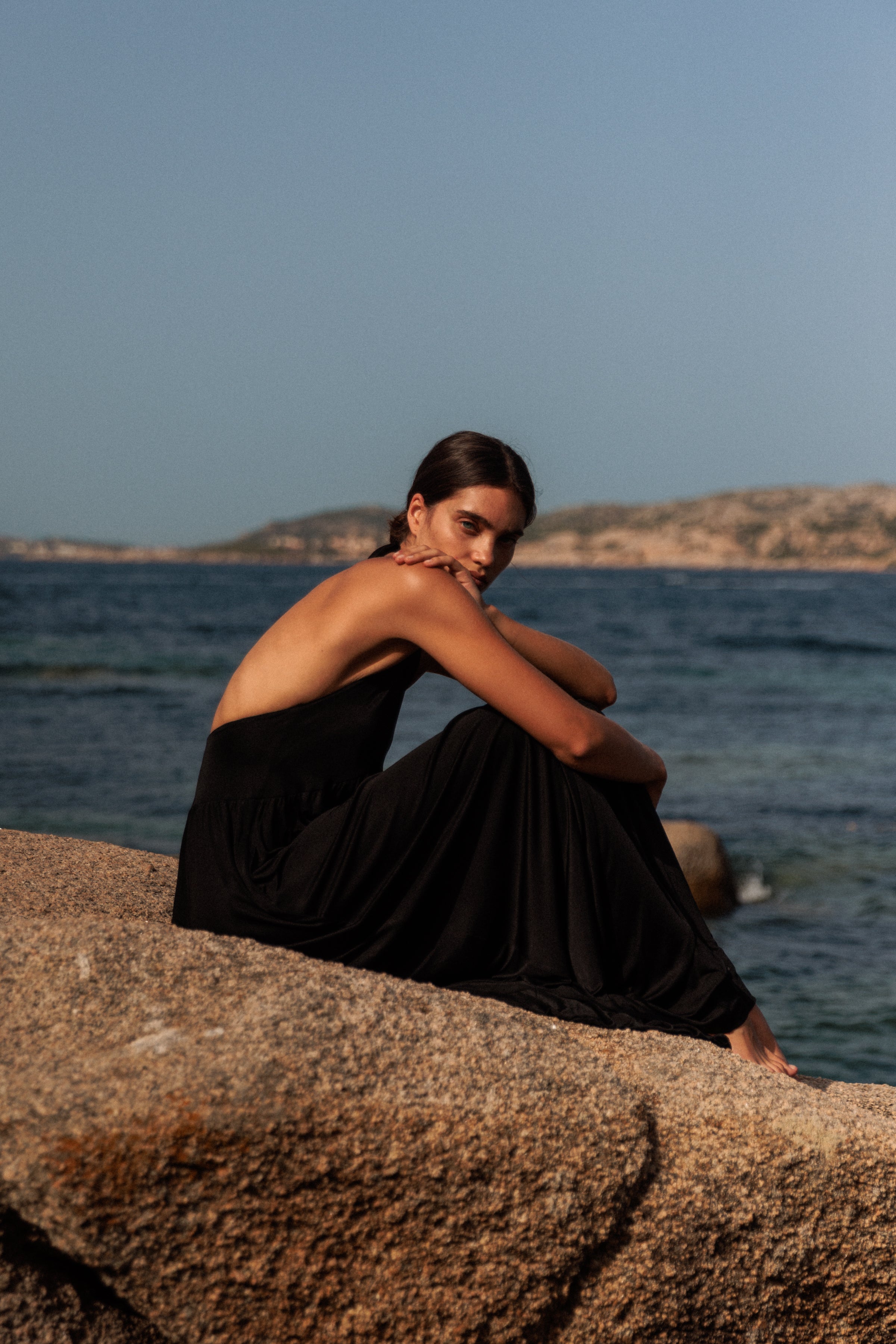 The woman is sitting on a large rock by the sea with knees drawn up and arms resting on them, looking out towards the water, wearing an Anna Halter black dress by Catherine Gee. The background features a calm ocean with distant rocky islands under a clear blue sky capturing a serene and contemplative moment by the seaside.