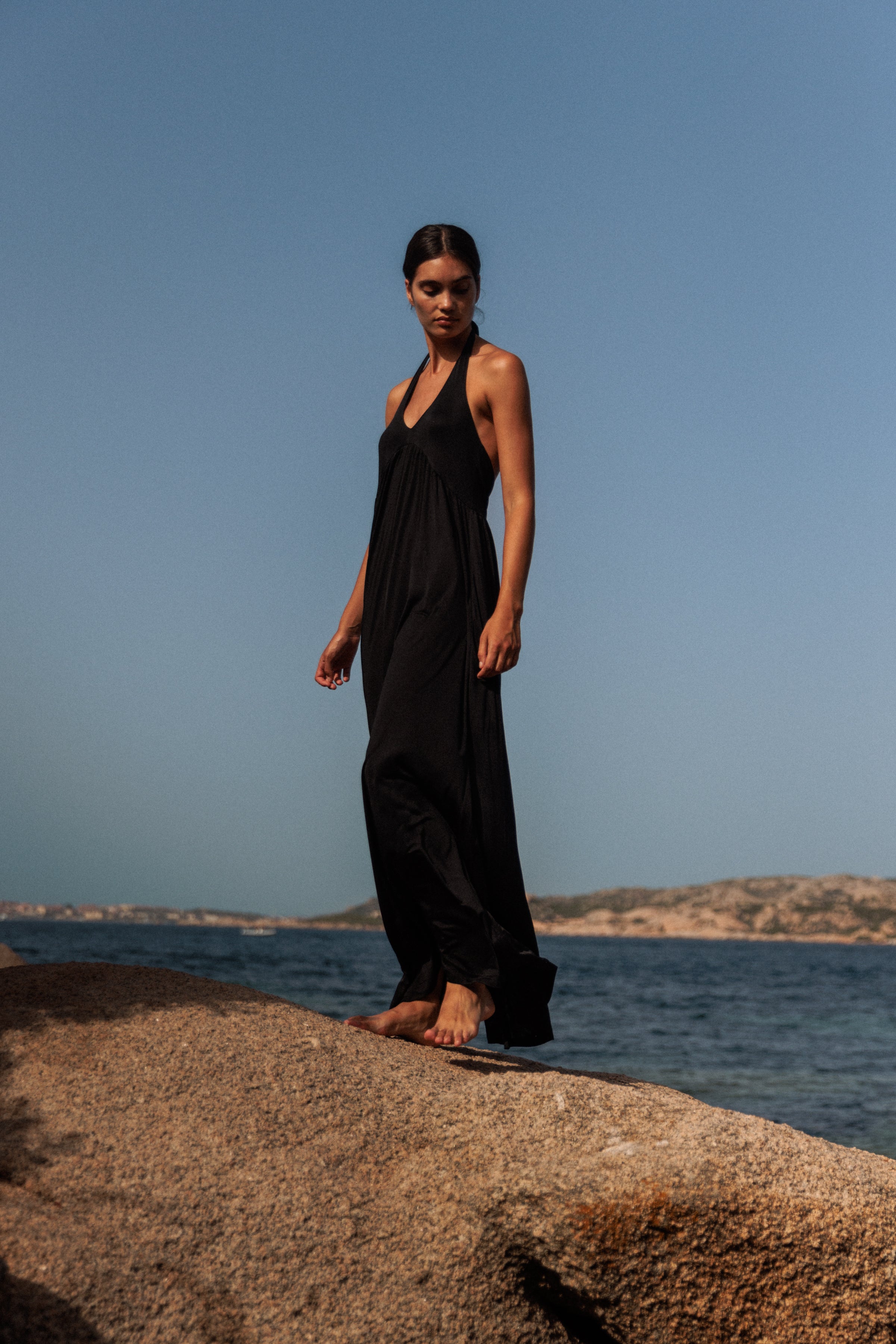 A woman standing on a large rock by the sea, wearing a long, Anna Halter black dress by Catherine Gee. The background features a clear blue sky and distant rocky hills across the water highlighting the elegance and flow of the dress in a natural, serene setting.