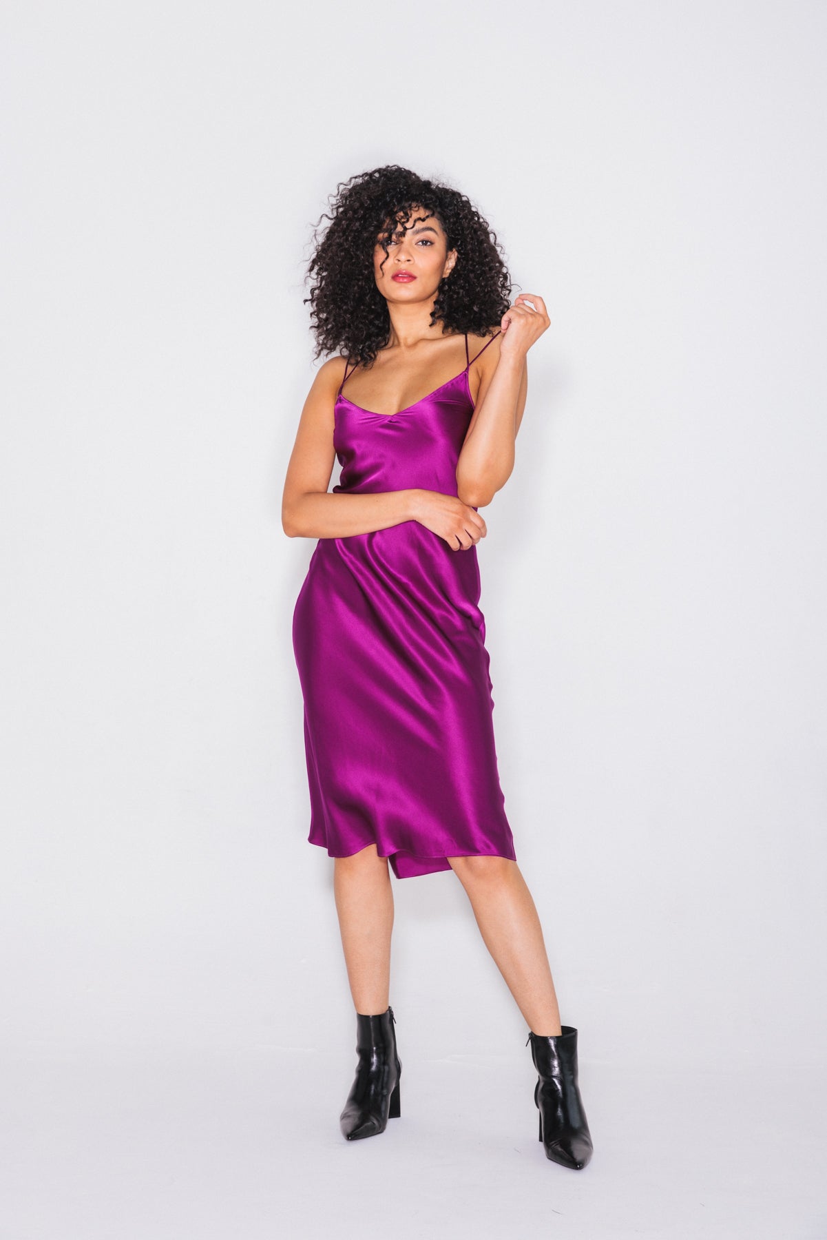 A woman wearing a Catherine Gee Emma Electric Orchid Slip Dress paired with black ankle boots, adds a stylish contrast to the dress. The dress is a vibrant purple color, made from a silky, satin-like fabric that drapes elegantly features thin spaghetti straps, and a fitted silhouette that falls to mid-calf length. The plain white background highlights the dress's color and texture.