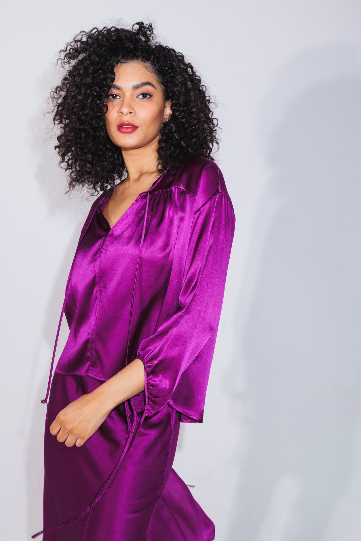 A woman wearing the Catherine Gee Frances Electric Orchid Blouse. The blouse is a vibrant, silky, and shiny purple color with a loose, flowing fit, and features a V-neckline and long sleeves with a slight puff at the shoulders. The fabric appears smooth and luxurious, reflecting light to enhance its rich hue. She also wears a matching skirt, creating a cohesive and elegant outfit.