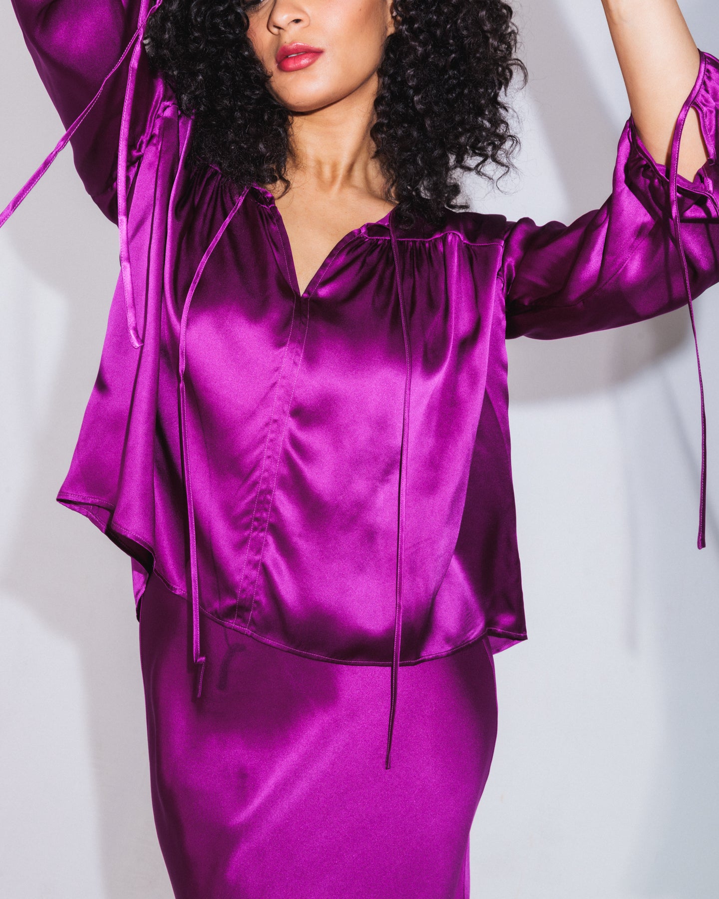 A woman wearing a Catherine Gee Frances Electric Orchid blouse, made from shiny, satin-like fabric in a vibrant purple color, has a loose fit, a V-neckline, and long sleeves with ties at the cuffs. The gathered details at the shoulders add elegance paired with a matching purple skirt, this coordinated outfit stands out for its bold color and luxurious texture, making it both stylish and eye-catching.