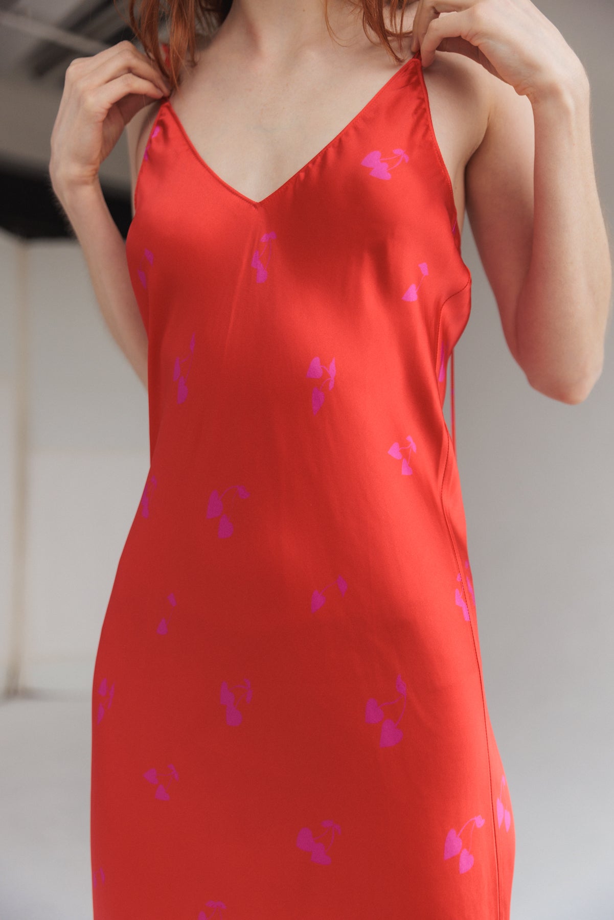 A woman adjusting the straps wearing a Catherine Gee Emma Cherry Heart Slip Dress. This vibrant red dress features a playful pattern of small purple hearts. It has a V-neckline and thin shoulder straps, offering a delicate and elegant look. The fabric appears smooth and silky, draping beautifully over the body highlighting the dress's perfect fit and sleek design.