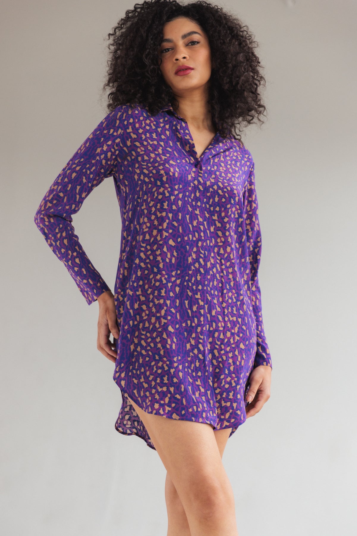 A woman wearing a Catherine Gee Daria Silk Purple Leopard dress is a long-sleeved, button-up shirt dress with a high-low hemline, features a vibrant purple color with an abstract leopard print pattern in a contrasting color. The fabric appears to be lightweight and flowy, giving it a relaxed yet stylish look. The dress is worn without any additional accessories, highlighting its bold pattern and elegant design.