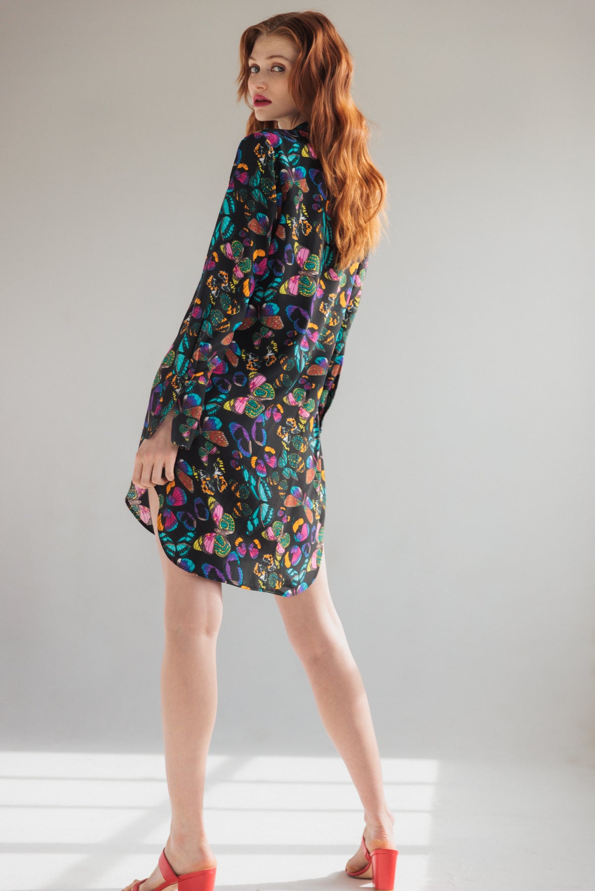 A woman wearing a Catherine Gee Daria Silk Shirt The Tracy dress features a multicolored butterfly print on a dark background, with long sleeves and a loose, flowing fit. Made from lightweight silk, the dress offers a luxurious feel. She standing with her back slightly turned, wearing red open-toe sandals, and is in a well-lit room with a plain background, which highlights the dress's eye-catching design.