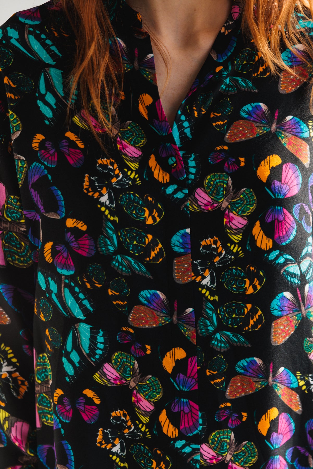 A close-up of the Catherine Gee Daria French Cuff Silk The Tracy blouse features a vibrant and colorful butterfly print on a black background. The butterflies are in various shades of blue, pink, orange, and green, creating a striking and eye-catching pattern. Made from smooth and luxurious silk, the blouse includes a collar and buttons down the front. The elegant and bold design makes it a standout piece for any wardrobe.