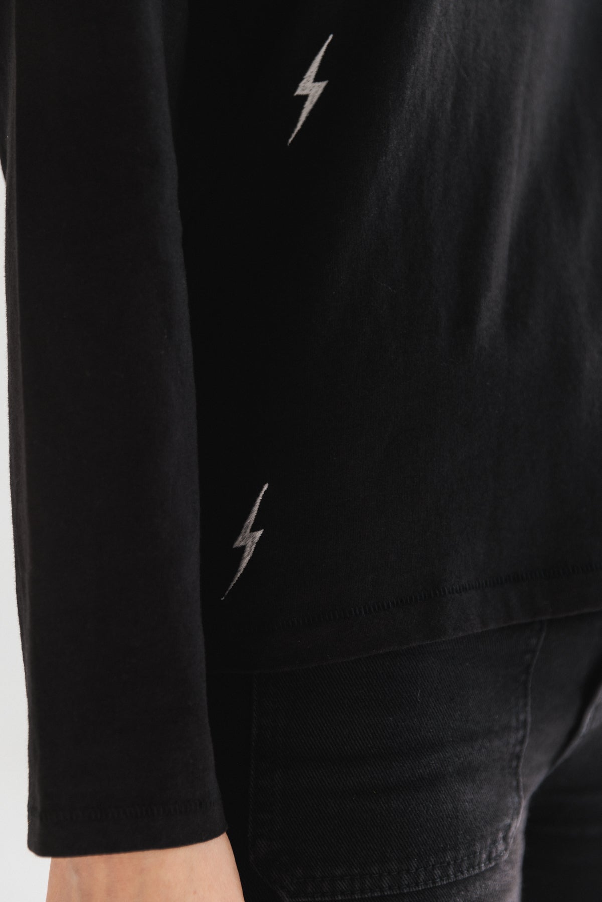A close-up of a woman wearing a black Catherine Gee Embroidered Cotton Long Sleeve Bolt T-Shirt features two small white lightning bolt symbols, one near the bottom of the sleeve and another near the bottom hem of the shirt. The focus is on the details of the clothing, especially the lightning bolt symbols, which add a distinctive and interesting element to the outfit.