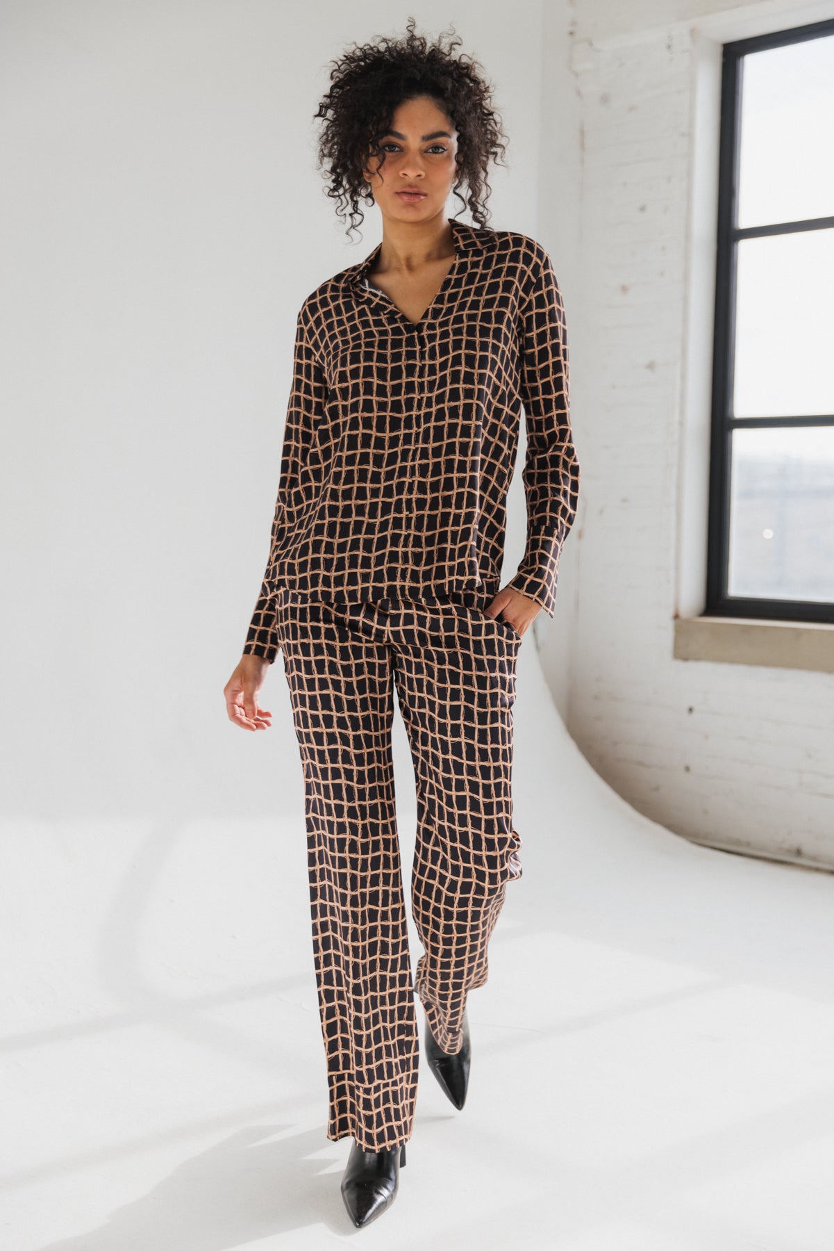 A woman wearing a Catherine Gee Daria French Cuff Silk Recoleta blouse features a geometric pattern with a grid-like design in black and beige colors and matching pants with the same pattern, creating a coordinated outfit. It has a relaxed fit with long sleeves and a v-neckline. The setting appears to be a bright, minimalist room with a large window, enhancing the overall elegance of the clothing.