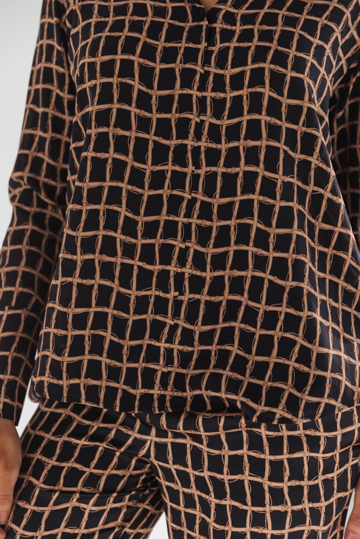 A close-up view of a woman wearing a Catherine Gee Daria French Cuff Silk Recoleta blouse features a distinctive grid pattern with beige lines on a black color background with matching pants sharing the same grid pattern, creating a coordinated and fashionable outfit. It has long sleeves, and a relaxed fit, making it both stylish and comfortable. The fabric appears to be smooth and silky, highlighting the luxurious nature of the material.