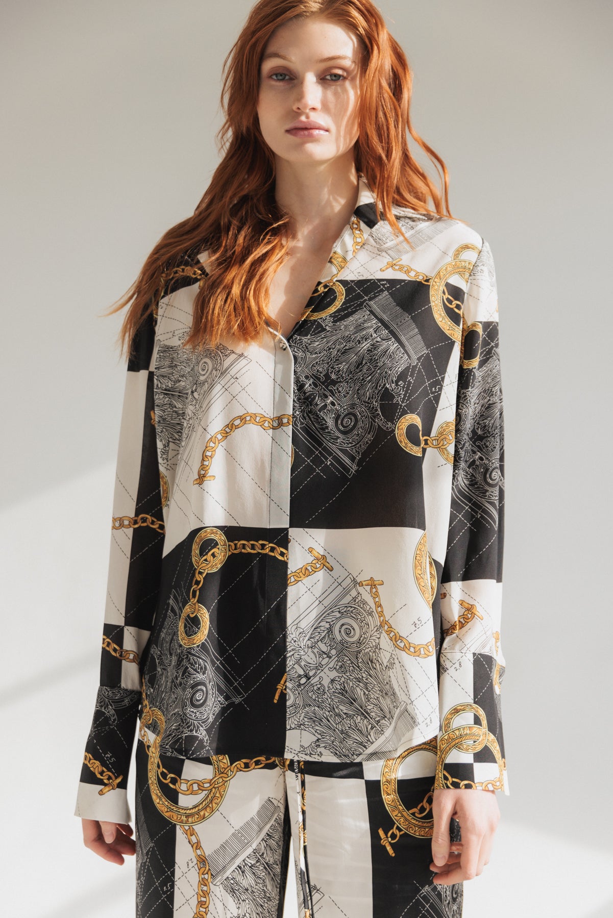 A woman wearing a Catherine Gee Daria French Cuff Silk - Athens Blouse, features a pattern of black and white sections with intricate designs and gold chain motifs. The blouse has a button-down front and long sleeves with French cuffs. The person has long, wavy hair and is standing against a plain background.