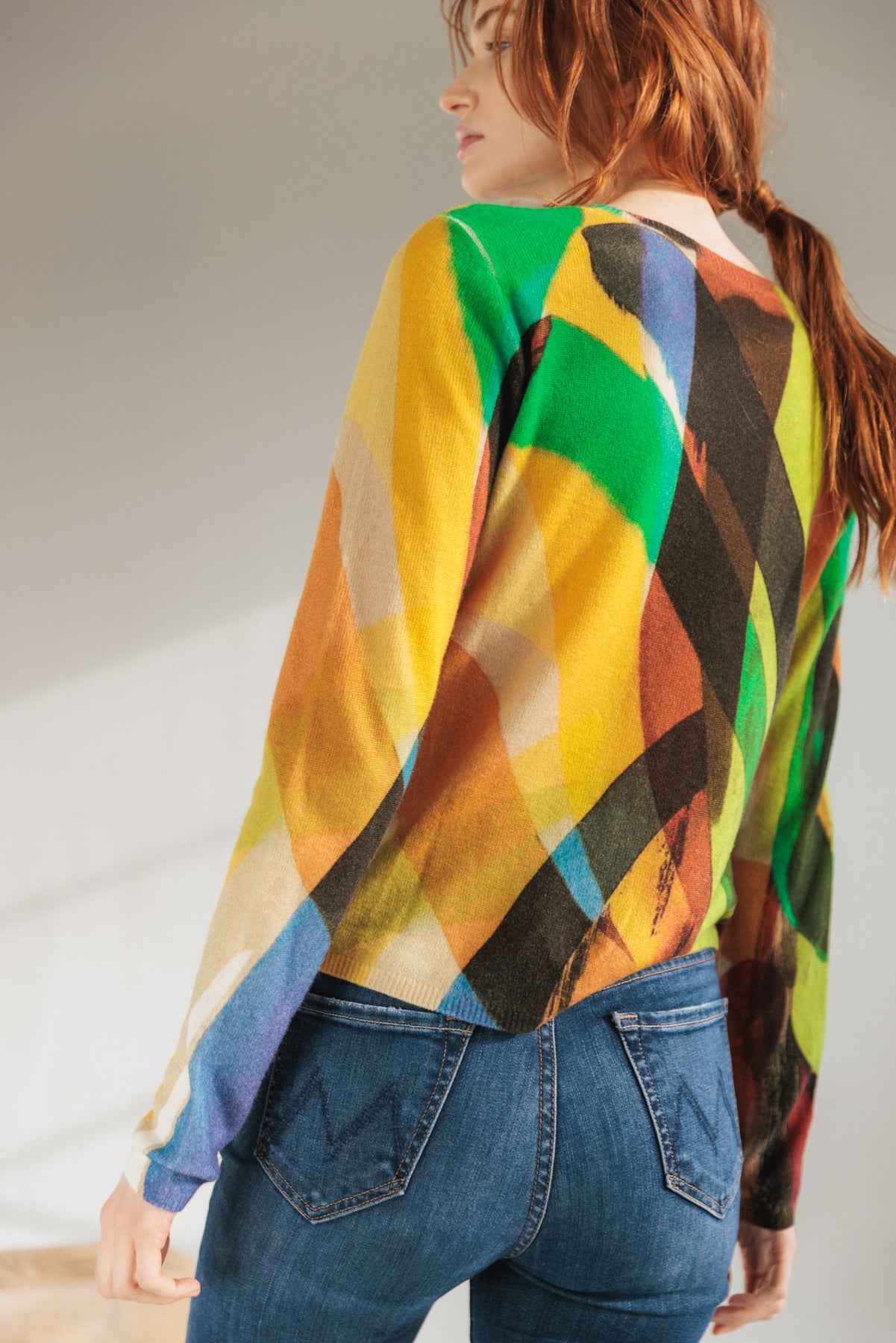 A woman is wearing a colorful Catherine Gee Crewneck Cashmere Sweater in the Stella Print design. The sweater features a vibrant, abstract pattern with various colors including green, red, yellow, blue, and black paired with blue jeans. The image is taken from behind, showing the back of the sweater and the jeans.