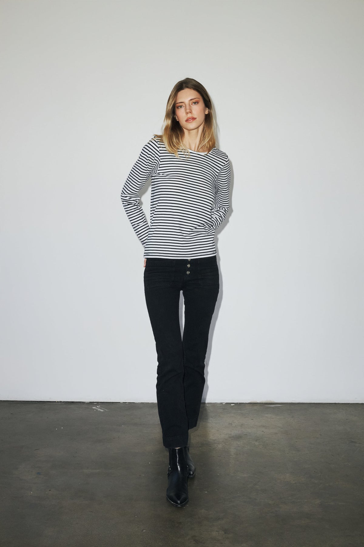 A woman is standing against a plain white background, wearing a black and white striped long-sleeve T-shirt by Catherine Gee. She is also wearing black pants and black ankle boots. The simplicity of the background focuses attention on the T-shirt's classic design and fit, making it the standout piece in the outfit.