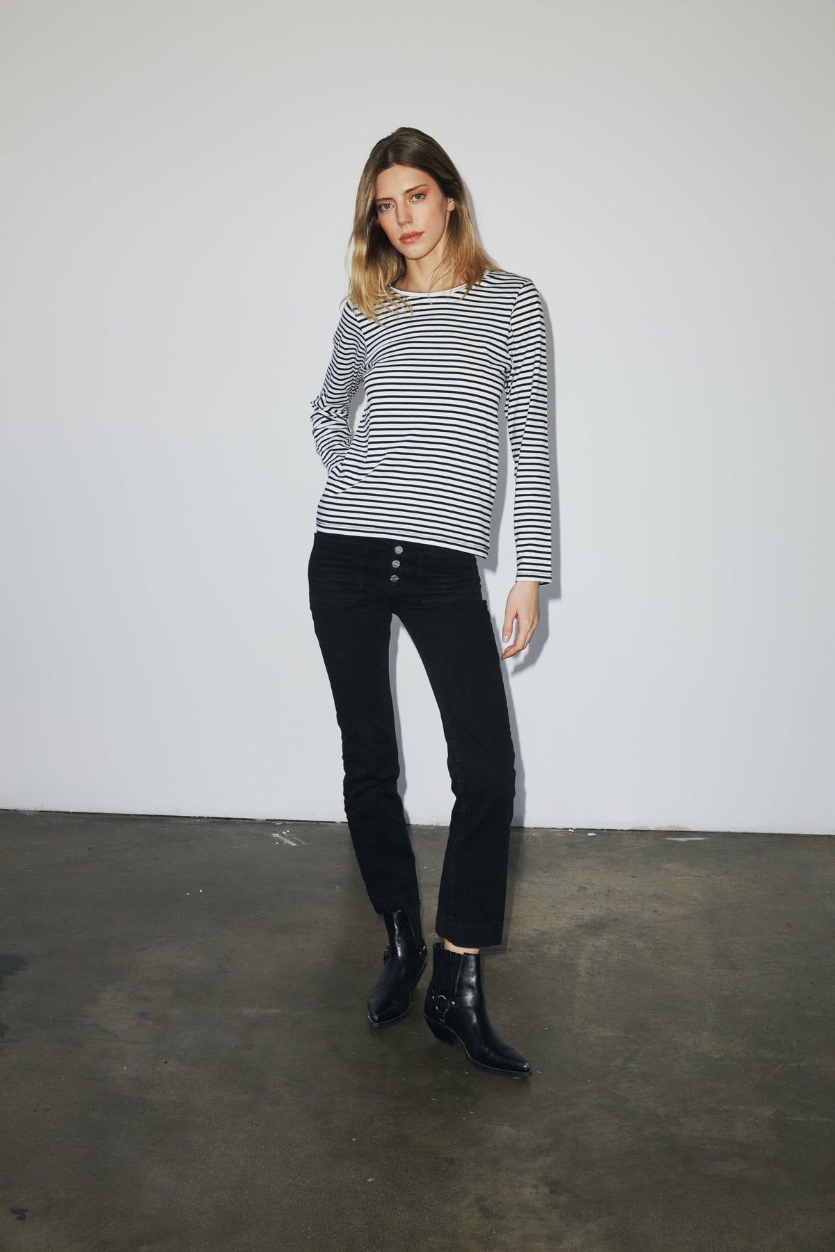 A woman is wearing a black and white striped long-sleeve T-shirt from Catherine Gee. The outfit is paired with black pants that have buttons on the front and black ankle boots.