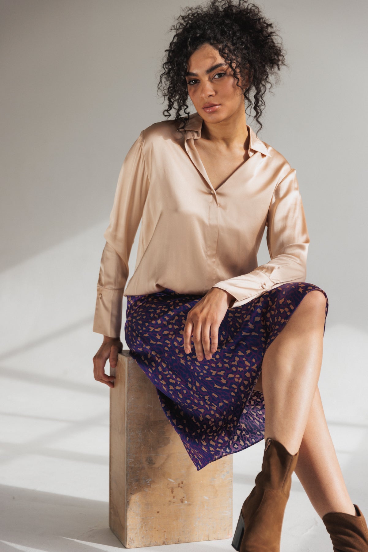 A woman is seated on a wooden block, wearing a Catherine Gee Daria French Cuff Silk-Champagne blouse paired with a purple skirt with a patterned design and brown ankle boots on a plain neutral background with a shade of light outside, shadowing the window frame highlighting its elegant and luxurious fabric and design.